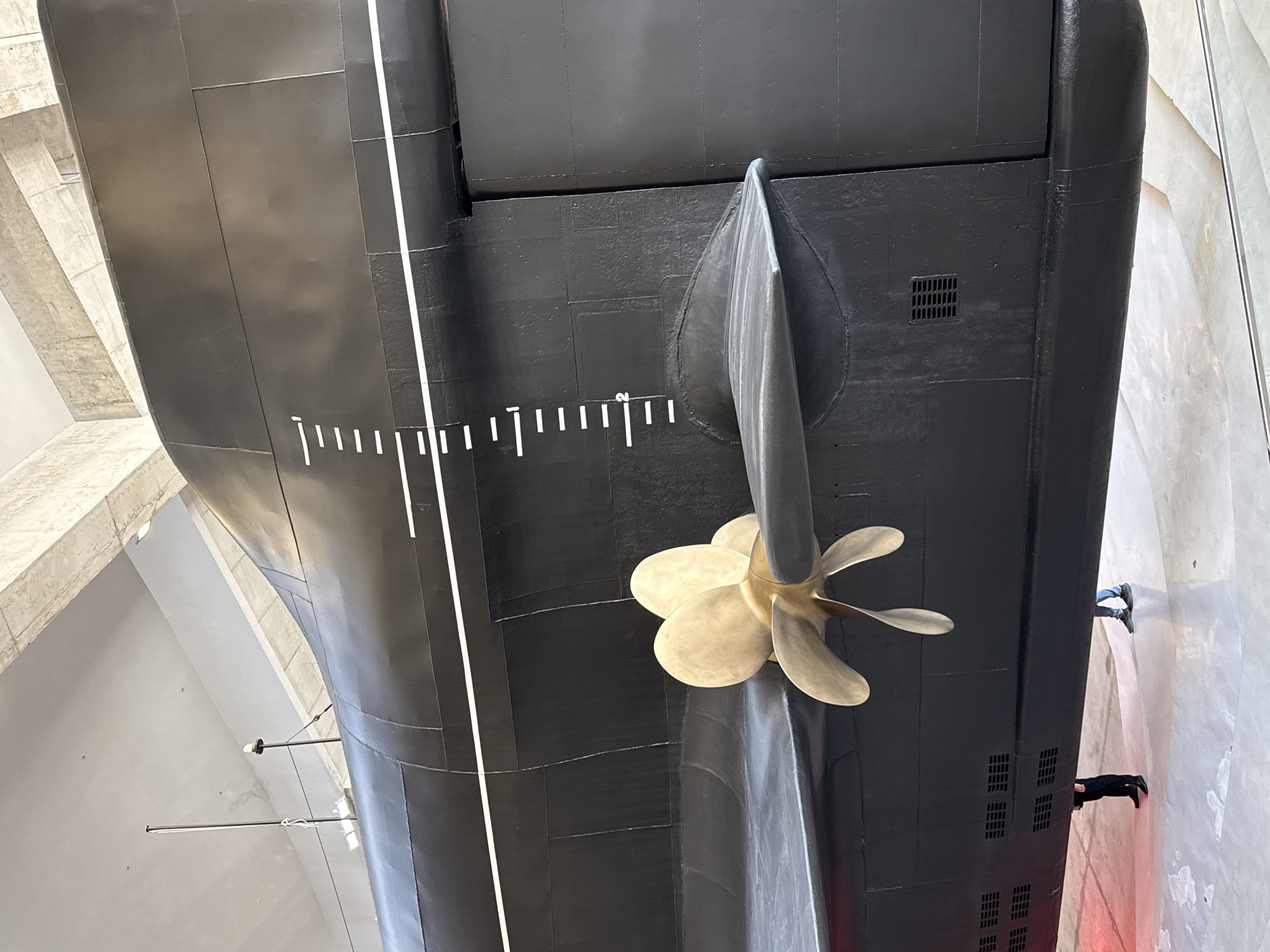 An extreme close-up, low-angle shot captures the stern of a black submarine, likely the S-189 submarine museum in Kronstadt, Russia. The focus is on the large, golden propeller, its three blades slightly blurred from a slow shutter speed. To the left of the propeller is a dark gray fin, also part of the submarine's stern. The submarine's hull is painted matte black, segmented by visible panel lines. A white line runs horizontally across the hull, and a vertical scale with white markings, indicating depth, is also present. In the lower portion of the image, the feet and legs of two individuals are visible, standing on a light-colored floor. One person, wearing dark trousers and black shoes, stands to the left. Another person, in blue jeans and dark shoes, stands to the right. The background consists of the submarine's superstructure and industrial-looking concrete ceiling elements, illuminated by overhead lighting.
