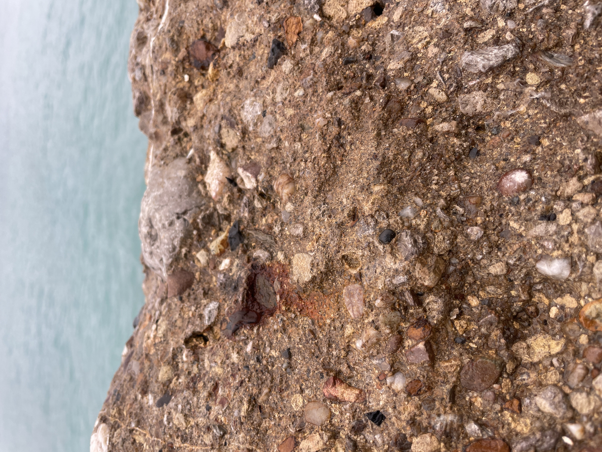 A close-up shot of a concrete wall speckled with small pebbles and stones of varying colors (brown, white, black, and reddish) and sizes, bound by a coarse, sandy matrix. A dark, almost blood-like stain is visible near the center-left of the frame. The left edge of the wall is ragged and abuts a flat, blue-gray surface, possibly water.
While it's not possible to confirm that the image was taken in Olginka, Russia from the image alone, a forensic description focuses on observable details only, not speculative conclusions.