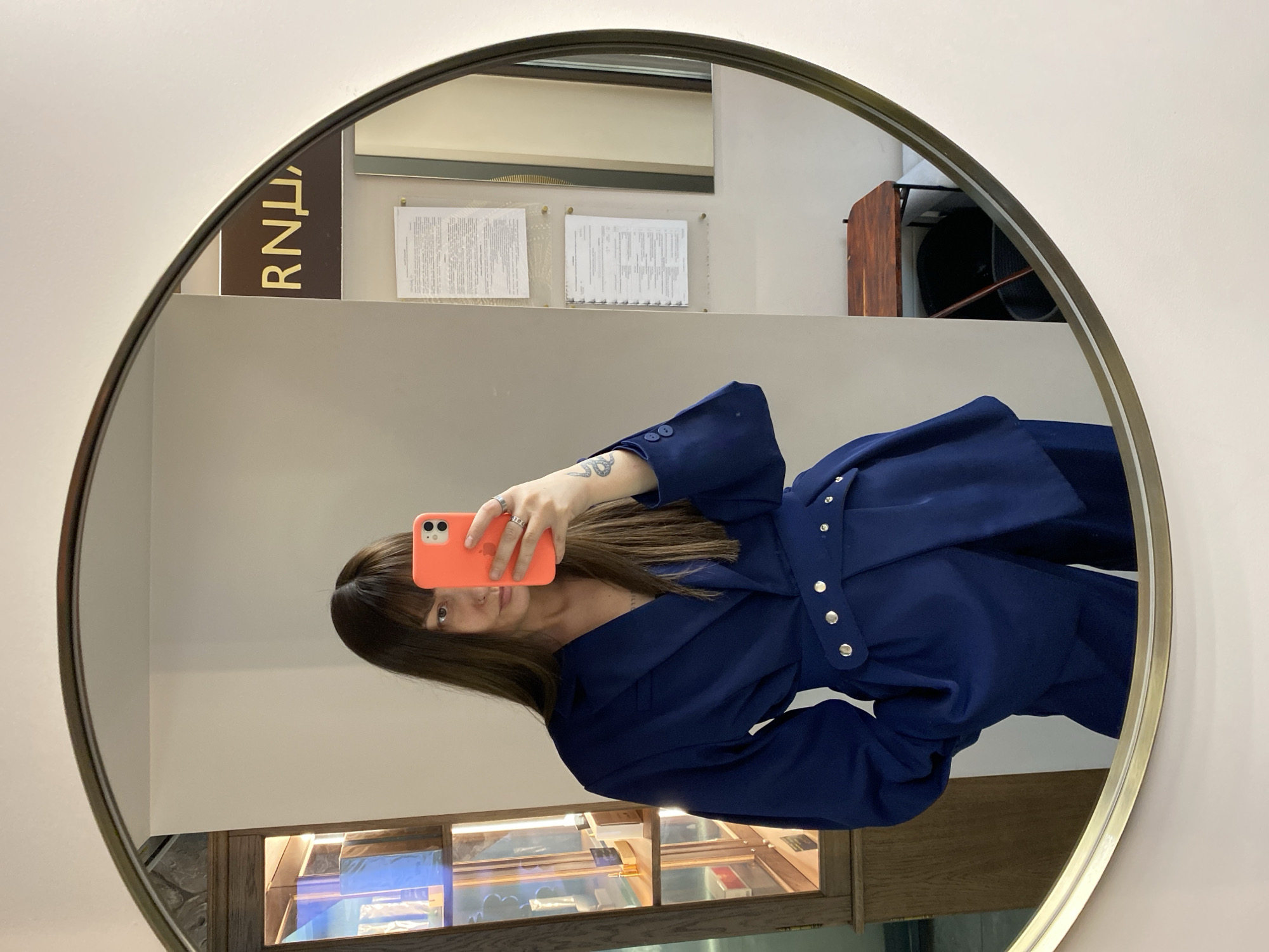 In Votkinsk, Russia, a woman takes a selfie in a round mirror. She holds an orange smartphone in front of her face, partially obscuring her features. The woman has long, brown hair with bangs and wears a dark blue belted jacket. Her hand with rings is visible. Reflected in the mirror, behind her, are documents on a wall, display cases, a brown cabinet and an "RNU" sign. The wall surrounding the mirror is white.
