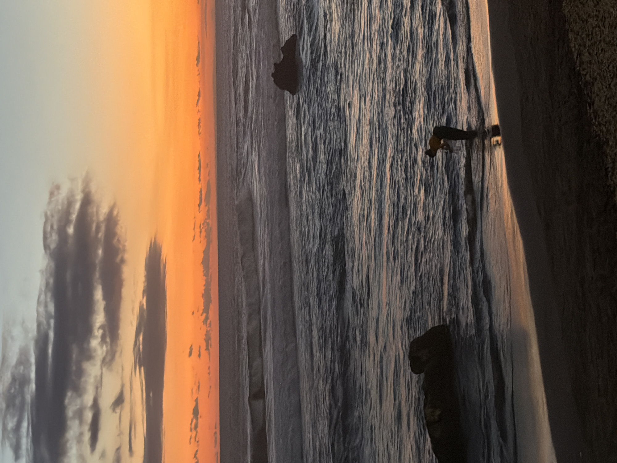 The image depicts a serene sunset over the ocean in North Kuta, Indonesia. The sky is ablaze with hues of orange and yellow, transitioning to a pale blue near the horizon. Wisps of grey clouds are scattered across the upper portion of the sky. In the foreground, a person, silhouetted against the water, stands at the edge of the sea, appearing to be interacting with the shallow waves. The water itself is a deep blue-grey, with white-capped waves rolling towards the shore. Two large rocks are visible in the water, one closer to the foreground on the left, and another further out on the right. The beach in the foreground is dark and sandy, reflecting the warm colors of the sunset. The overall impression is one of tranquility and natural beauty.