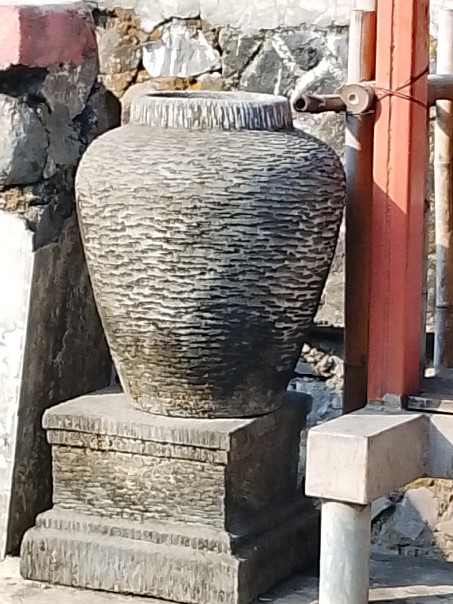 A tall, textured ceramic urn with a wide mouth sits atop a rectangular stone pedestal. The urn is dark gray and has horizontal ridges encircling its body. The pedestal is also stone, with visible layers and texture. Behind the urn, a rough stone wall with some red paint on the upper left is visible. To the right of the urn, a red vertical beam stands next to a metal railing. A small, hook-like object is attached to the beam with a wire. The scene appears to be outdoors, possibly in an urban environment in Kediri City, Indonesia, given the presence of stone structures and railings. The lighting suggests a sunny day.