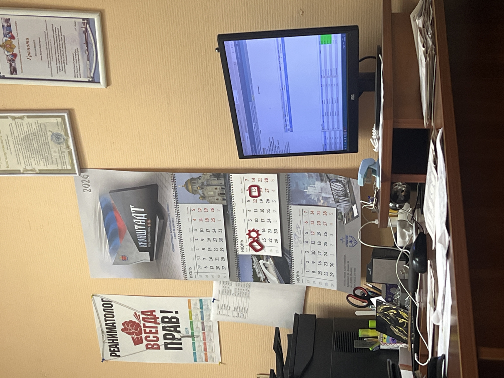 The image depicts a close-up view of a desk in what appears to be an office environment. On the wall, a poster with Russian text, "ВСЕГДА ПРАВ!", which translates to "ALWAYS RIGHT!", is prominently displayed. To the right of this poster, a calendar for 2026 is visible, featuring an image of Kronstadt, Russia, a port city on Kotlin Island in the Gulf of Finland. The calendar pages for June, July, and a portion of August are visible, with some dates marked. Further to the right on the wall, two framed documents, likely certificates or awards, are hung.
The desk itself is cluttered with office supplies. A computer monitor displays a spreadsheet-like interface. To the left of the monitor is a printer. Various items like pens, scissors, a stapler, a computer mouse, and stacks of papers are scattered across the desk surface. The overall impression is of a functional, albeit somewhat disorganized, workspace. The background wall is a light, textured yellow.
