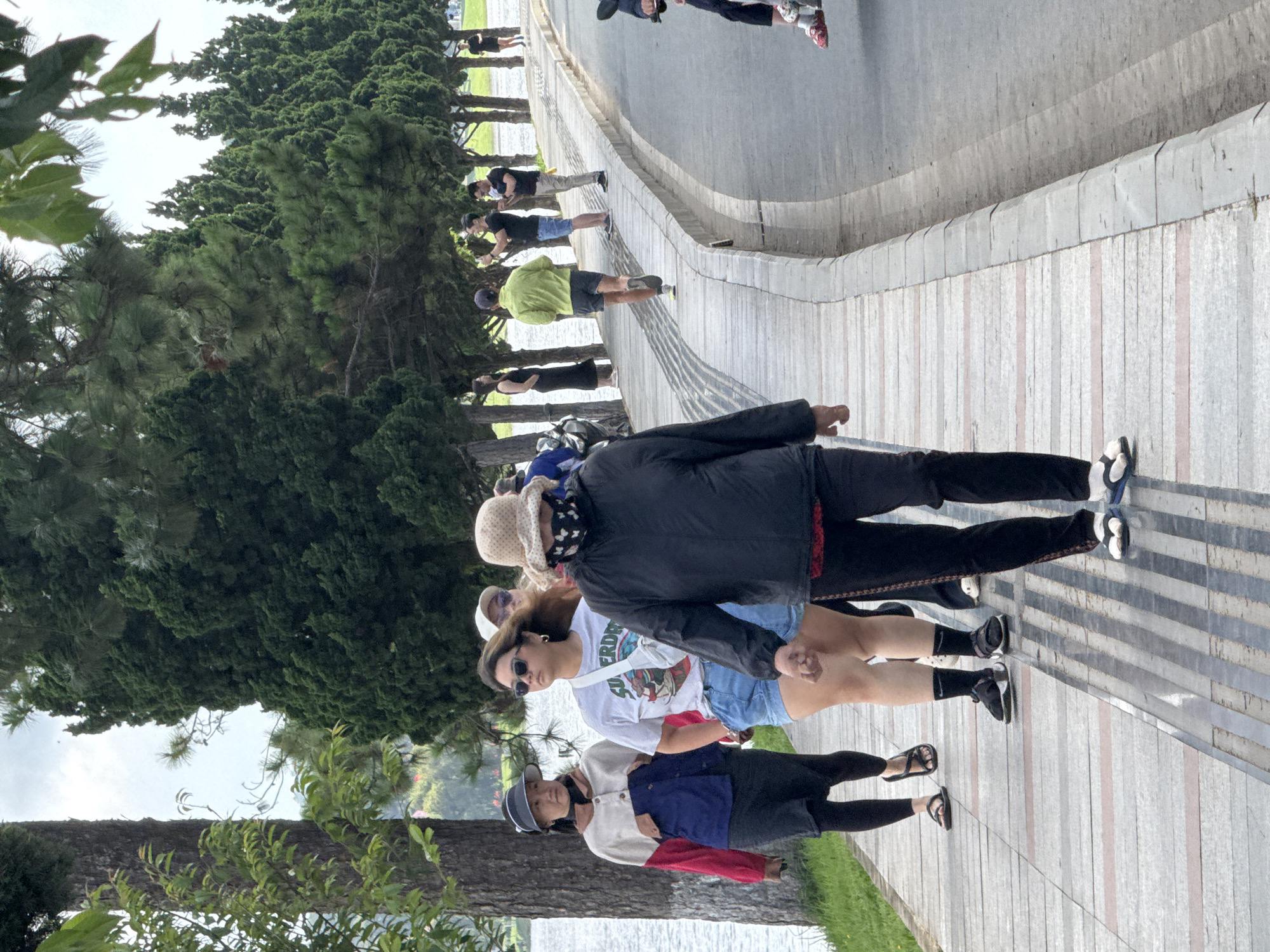 This image captures a daytime scene along the paved pedestrian pathway of Xuan Huong Lake in Da Lat, Vietnam, under an overcast sky. The pathway, constructed from grey and reddish-brown striped pavers, runs parallel to a tranquil body of water (the lake) on the left and a grey asphalt road on the right.
In the foreground, three individuals are walking towards the background on the pathway. From left to right:
1. An adult woman wears a dark visor, a long-sleeved jacket with red, white, and dark blue sections, a black skirt, black leggings, and black thong sandals. She looks slightly to her right.
2. A younger adult woman wears sunglasses, a light grey t-shirt with "SUPERDRY" branding, denim shorts, black athletic socks, and dark athletic sandals. A white cross-body bag is visible across her chest. She walks forward.
3. An adult woman is partially seen from the back, wearing a light-colored wide-brimmed hat, a patterned dark face mask, a black long-sleeved jacket, black pants with red trim, and white and blue flip-flops.
Further along the pathway in the midground, several other individuals are present. A person in a lime green athletic long-sleeve top and dark shorts is jogging away from the viewer. To their right, a man in a black t-shirt and blue shorts is holding a smartphone upwards, apparently capturing a photo or video. Next to him, another man in a black t-shirt and light-colored pants is looking at a smartphone. To the left of the jogger, a woman in a long black dress stands facing the lake.
The left side of the scene is dominated by a large tree trunk with light-colored bark patterns, situated between the pathway and the lake. Numerous tall, slender evergreen trees, resembling cypress or pine, line both sides of the pathway, extending into the background and forming a dense green canopy overhead. A concrete curb separates the pathway from the adjacent road. In the far background on the right, another individual in dark attire is standing near the trees, and the lower legs and red footwear of a person are partially visible on the extreme right edge, suggesting another presence, possibly on a small vehicle. The lake surface exhibits minimal ripples.