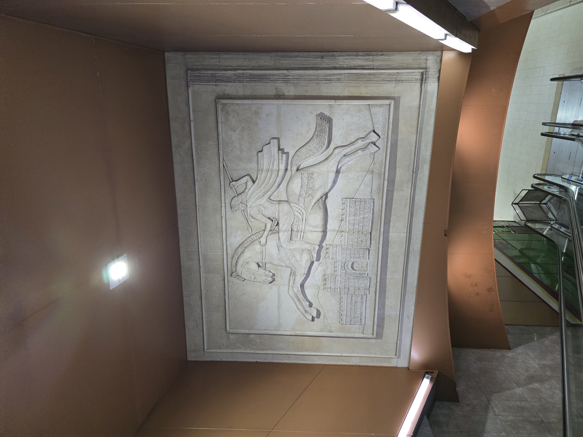 The image shows a bas-relief sculpture on a wall in a public space, likely a subway station in Sofia, Bulgaria. The sculpture depicts a winged horse and rider, possibly of historical or mythological significance, positioned above a stylized architectural element. The background wall is painted in a dark tan or brown. The bas-relief is off-white or gray, framed in a textured rectangle. To the right, a curved structural element is visible, also painted in tan, and an escalator leads upward. The overall lighting appears to be artificial, with at least one light fixture visible near the left edge of the image. The scene looks to be a modern transportation area with a mix of modern architecture and artistic elements.