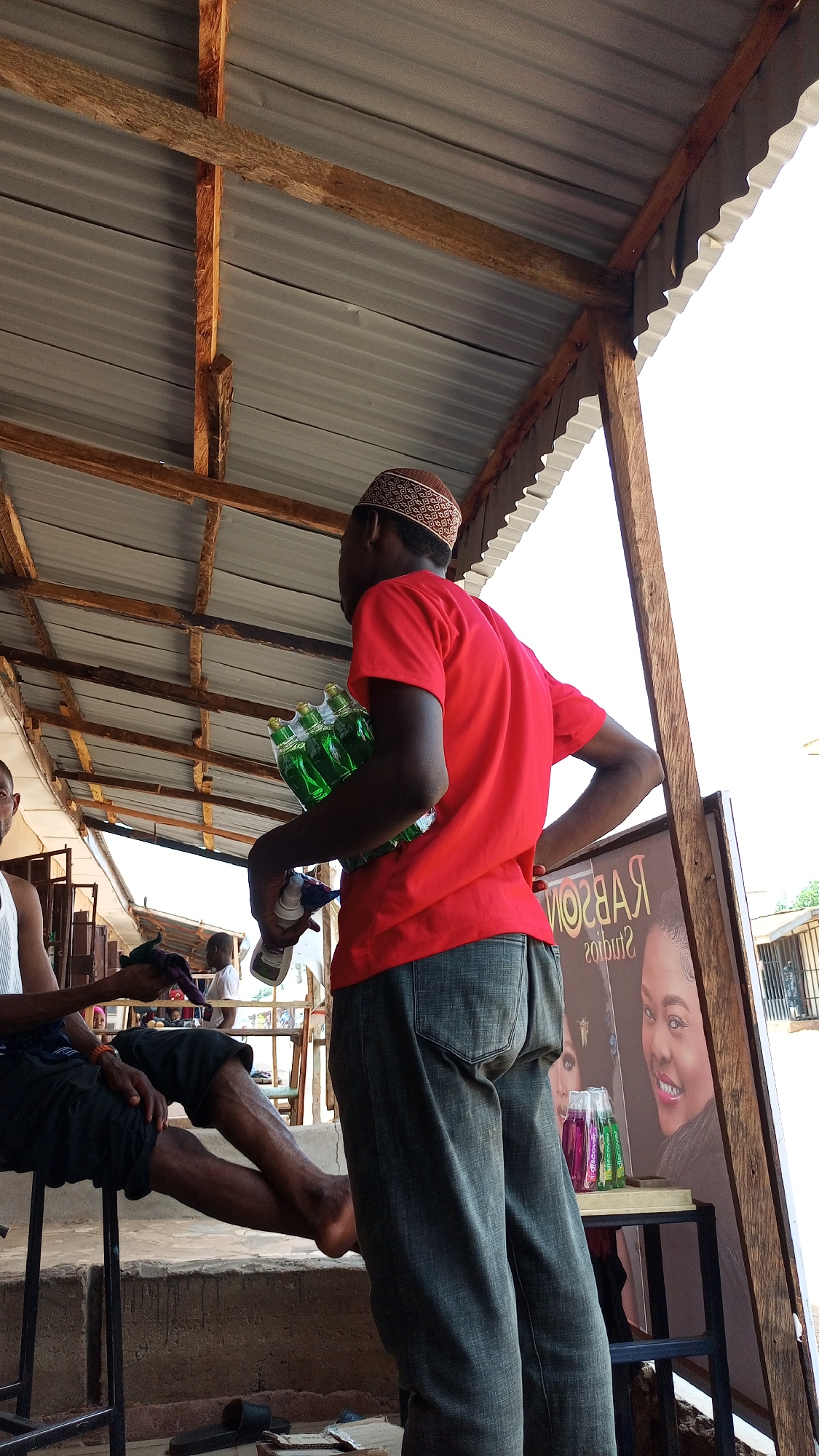 The image captures a scene in Sabon Tasha, Nigeria, under a corrugated metal roof supported by wooden beams. A man in a red t-shirt, dark jeans, and a patterned kufi is standing, holding a set of green bottled drinks and a white spray bottle. His back is to the viewer, and he appears to be looking to his left. Another man is seated on a metal stool, facing to the left, with one leg extended and bare feet visible. A third person can be seen sitting in the background, further to the left. A metal framed sign with images of women, and the text "RABSON studios," stands near the standing man. In the foreground, there's a step made of concrete, and various objects including a black sandal and what appears to be a piece of metal.