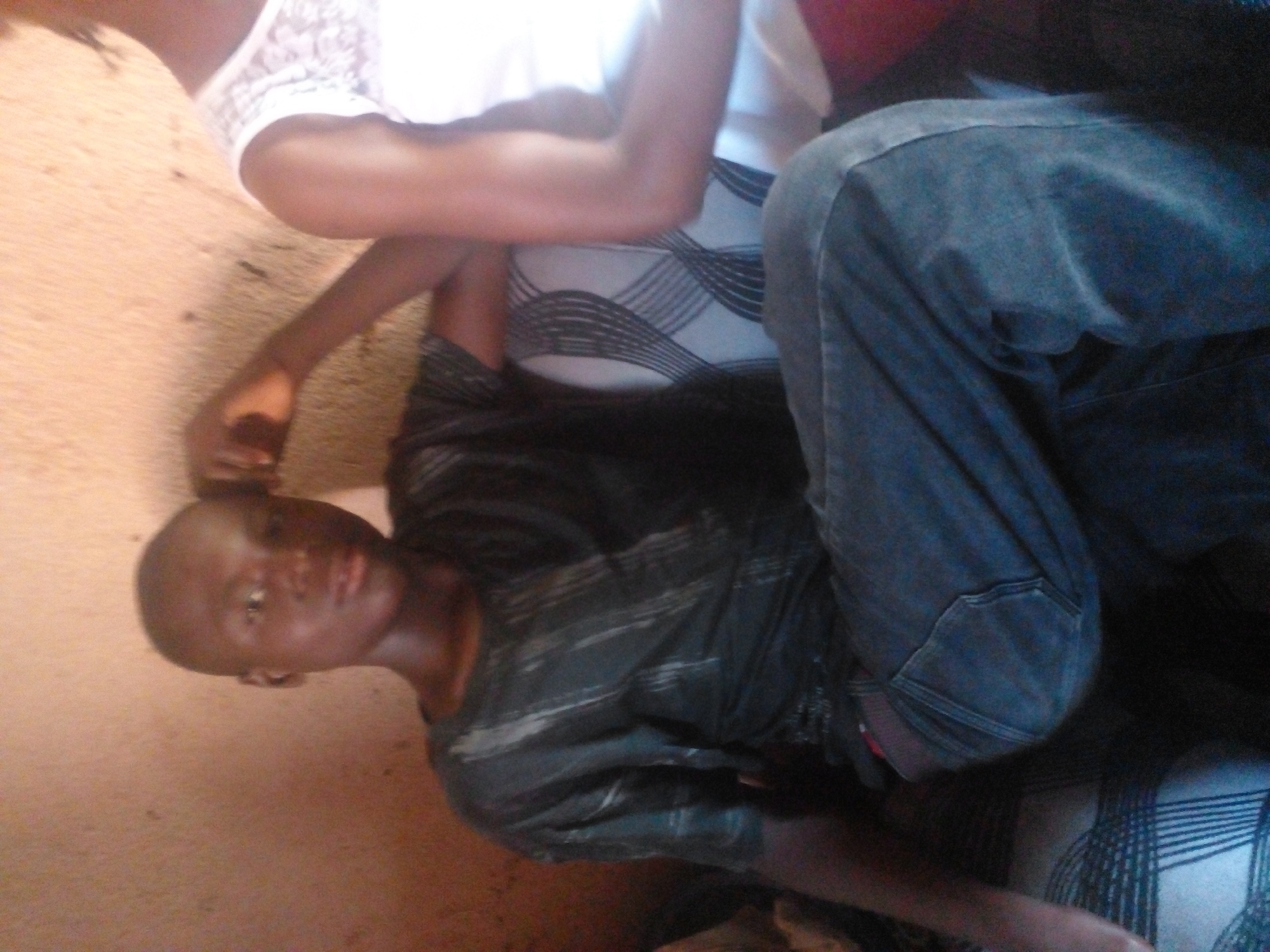 In an indoor setting with a plain, textured wall as a backdrop, a young Black male with a shaved head is seated on a patterned couch. He is wearing a dark, distressed t-shirt and blue jeans. His left arm is bent, and his hand rests on his forehead, suggesting a relaxed or pensive pose. To his right, another individual, partially visible, wears a white lace top. The lighting is warm and appears to be natural, casting shadows that contribute to the intimate atmosphere of the scene. The image has a slightly blurry quality, giving it a candid, unposed feel.