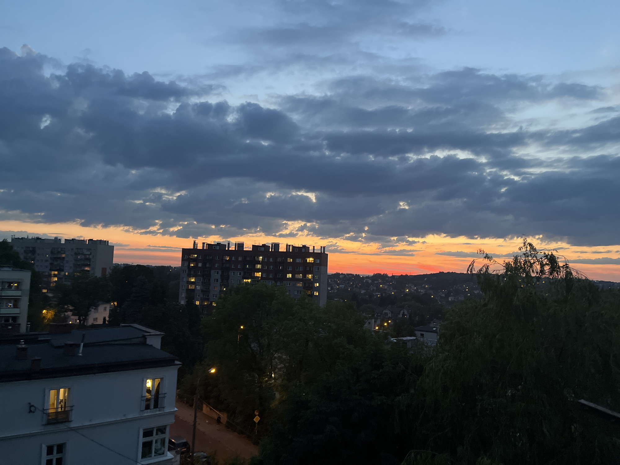 The image depicts an urban landscape at dusk. A prominent multi-story apartment building with illuminated windows stands in the middle ground. In the foreground, a white building with dark rooflines is visible, featuring lit windows and small balconies. The sky is a dramatic blend of dark clouds, with a vibrant orange and red glow concentrated along the horizon, indicative of a sunset. The city stretches into the distance, with numerous scattered lights suggesting residential areas. Lush green trees are interspersed throughout the scene, some obscuring parts of the buildings. The overall atmosphere is one of a city winding down for the evening under a colorful sky.