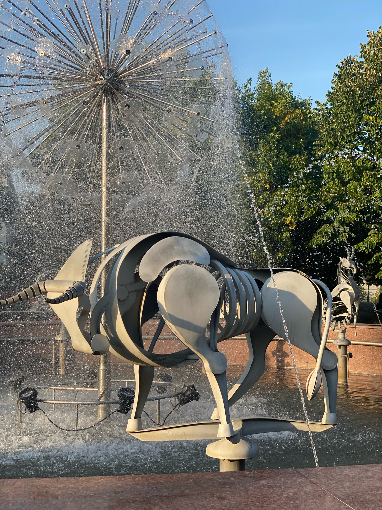 A metal sculpture of a bull skeleton stands in the foreground of a fountain. The sculpture is light gray and abstract, with curved metal plates forming the body and rib cage. The bull is facing left, with its head turned slightly towards the viewer. In the background, a spherical fountain sprays water upwards and outwards, creating a misty effect. The water droplets catch the sunlight, creating a glittering appearance. Behind the fountain, lush green trees are visible against a clear blue sky. The overall scene is peaceful and artistic. The bull sculpture is located in front of the fountain in the area around the National Opera and Ballet Theatre "Maria Bieșu" in Chișinău, Moldova.