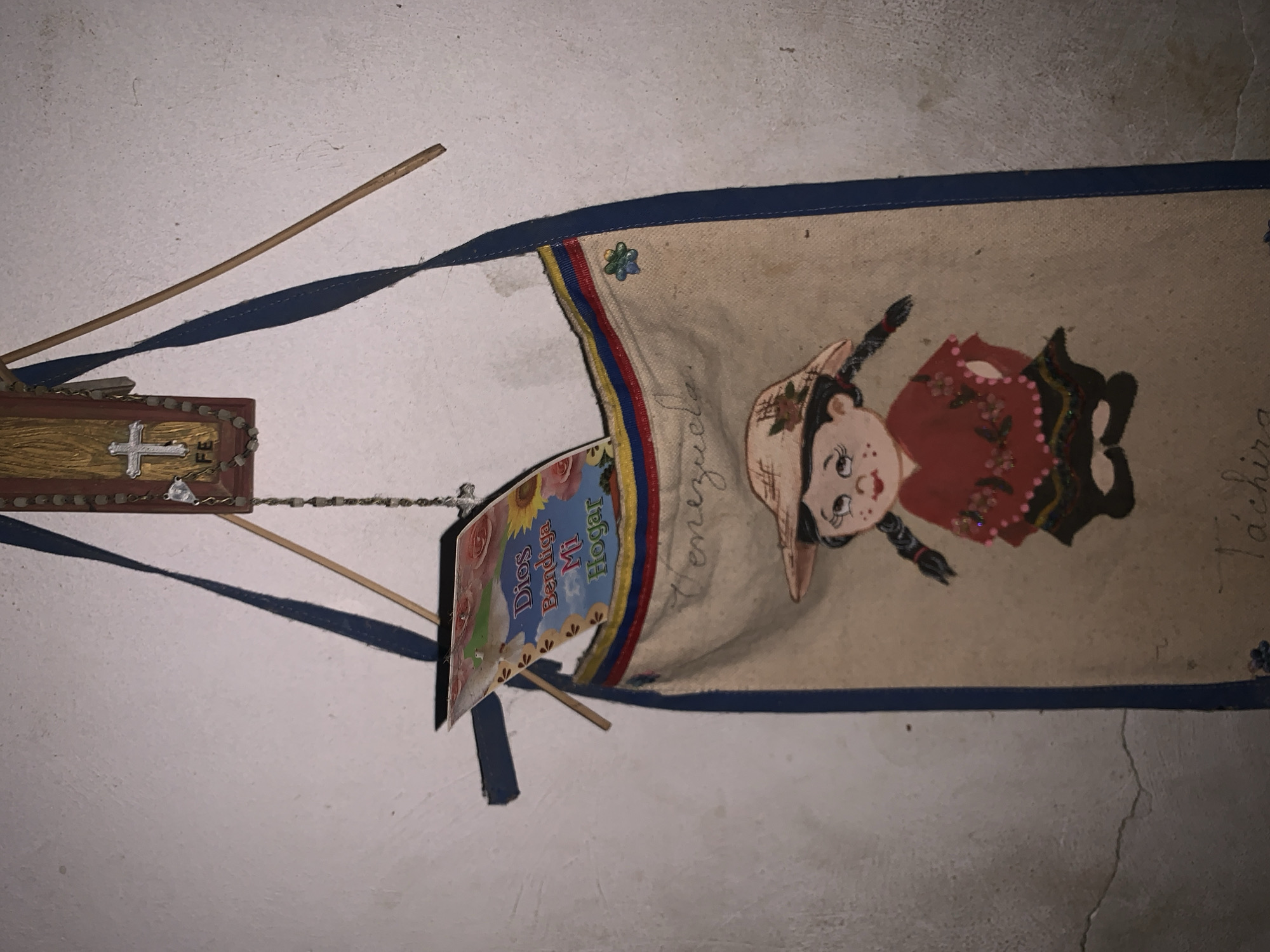 The image captures a collection of decorative and religious items displayed on a textured white wall, likely indoors. The main subject is a fabric banner, oriented vertically, featuring a cartoon-style illustration of a girl dressed in traditional attire, including a hat, and what appears to be a red dress with elaborate details. The banner has a blue trim and a vertical stripe in yellow, blue, and red, reminiscent of the Venezuelan flag, along with a small flower emblem. Text on the banner reads "Venezuela" and "Táchira," indicating a connection to the Táchira state in Venezuela.
To the left of the banner, a small wooden plaque or frame hangs. It is decorated with a wood-grain pattern and features a white cross. Next to the cross, the letters "FE" (Spanish for "Faith") are visible. A string of rosary beads extends from this plaque, connecting to a smaller, colorful paper flag. This small flag is adorned with floral and sunflower designs and bears the Spanish inscription "Dios Bendiga Mi Hogar" (God Bless My Home).
Two thin wooden sticks, possibly incense sticks or part of the hanging structure, are visible, casting long, defined shadows on the wall. The lighting suggests an artificial source or strong directional light. The arrangement of these items conveys a sense of domesticity, religious devotion, and national pride rooted in Venezuela, specifically Santa Ana.