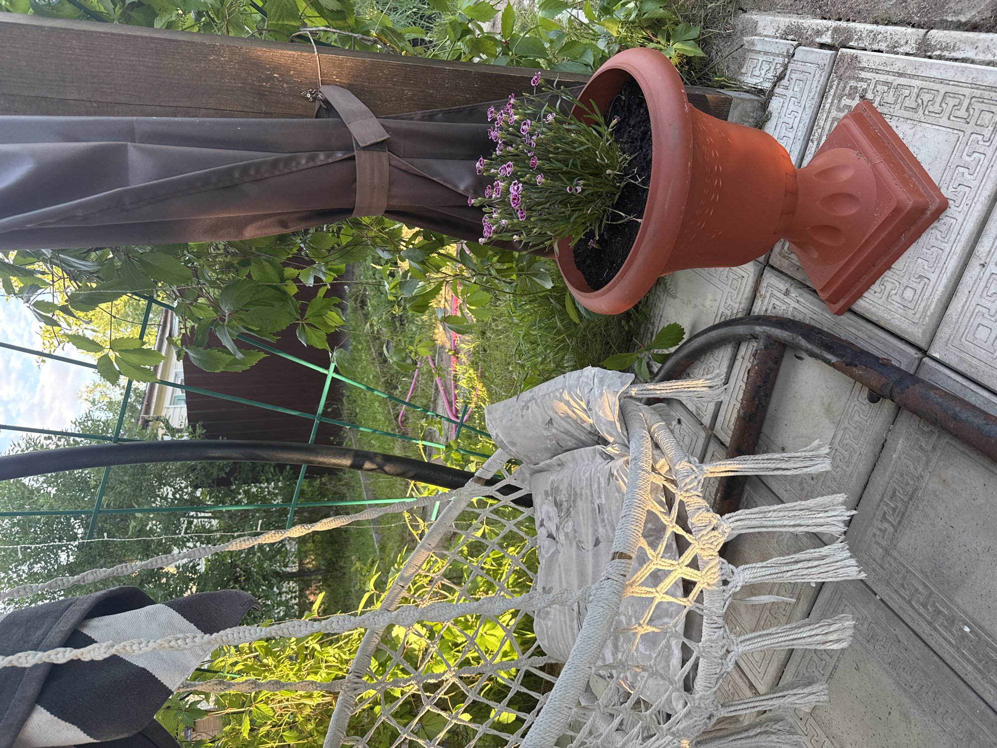 In the city of Tmz Snt, Russia, a serene outdoor setting is captured. The scene features a macrame hammock chair, adorned with a patterned pillow, suspended from a metal frame. Adjacent to the chair, a terracotta planter overflows with delicate pink flowers. The ground beneath is paved with decorative square tiles, and the backdrop is a lush garden environment with green foliage and trees. The lighting suggests it is either early morning or late afternoon, with warm sunlight casting long shadows.