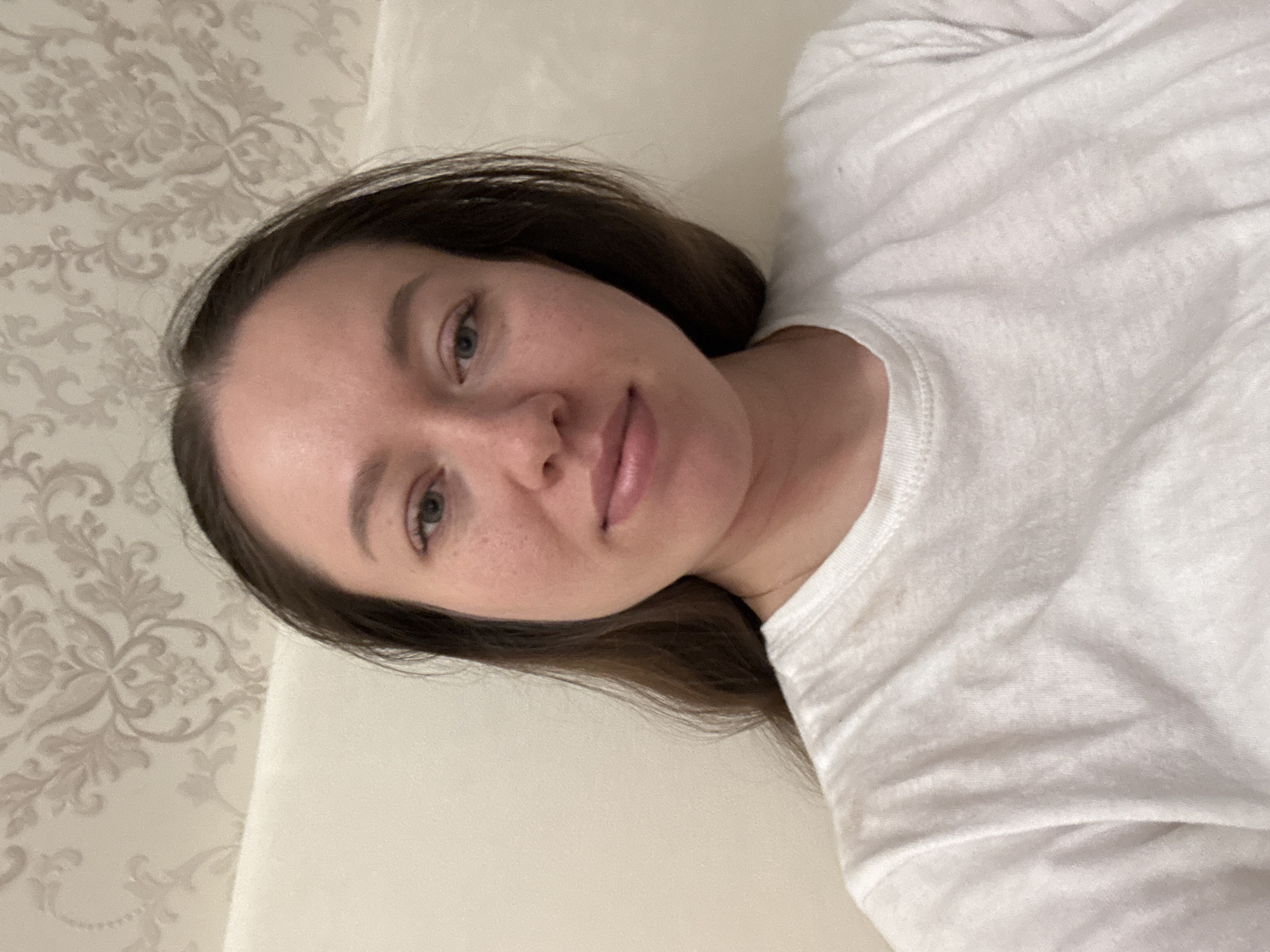A woman with brown hair and fair skin is lying down, possibly on a bed or sofa. She is wearing a white shirt. The background includes a patterned wallpaper on the left and a plain white surface (likely a bed or wall) behind her. The photo appears to have been taken indoors, with the subject looking directly at the camera. There is no definitive evidence to confirm this was taken in or near Ферма, Russia.