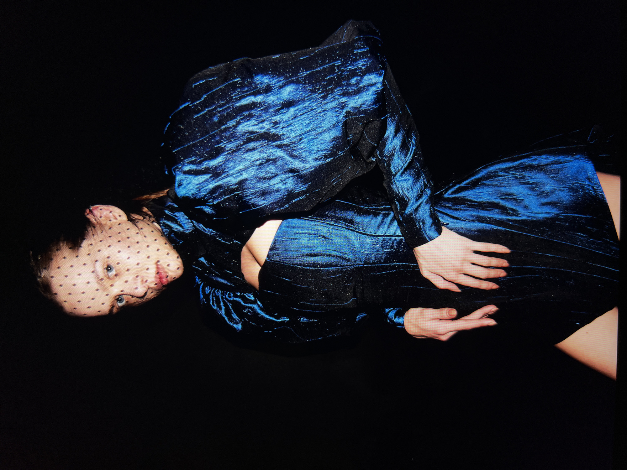 A woman with light skin and light blue eyes is posing against a black background. She is wearing a black and metallic blue dress with puffed sleeves and a veiled headpiece dotted with black polka dots. Her hands are positioned in front of her torso, one with fingers slightly bent and the other with fingers spread. The lighting is dramatic, highlighting the texture and sheen of the dress. The location is not specified.