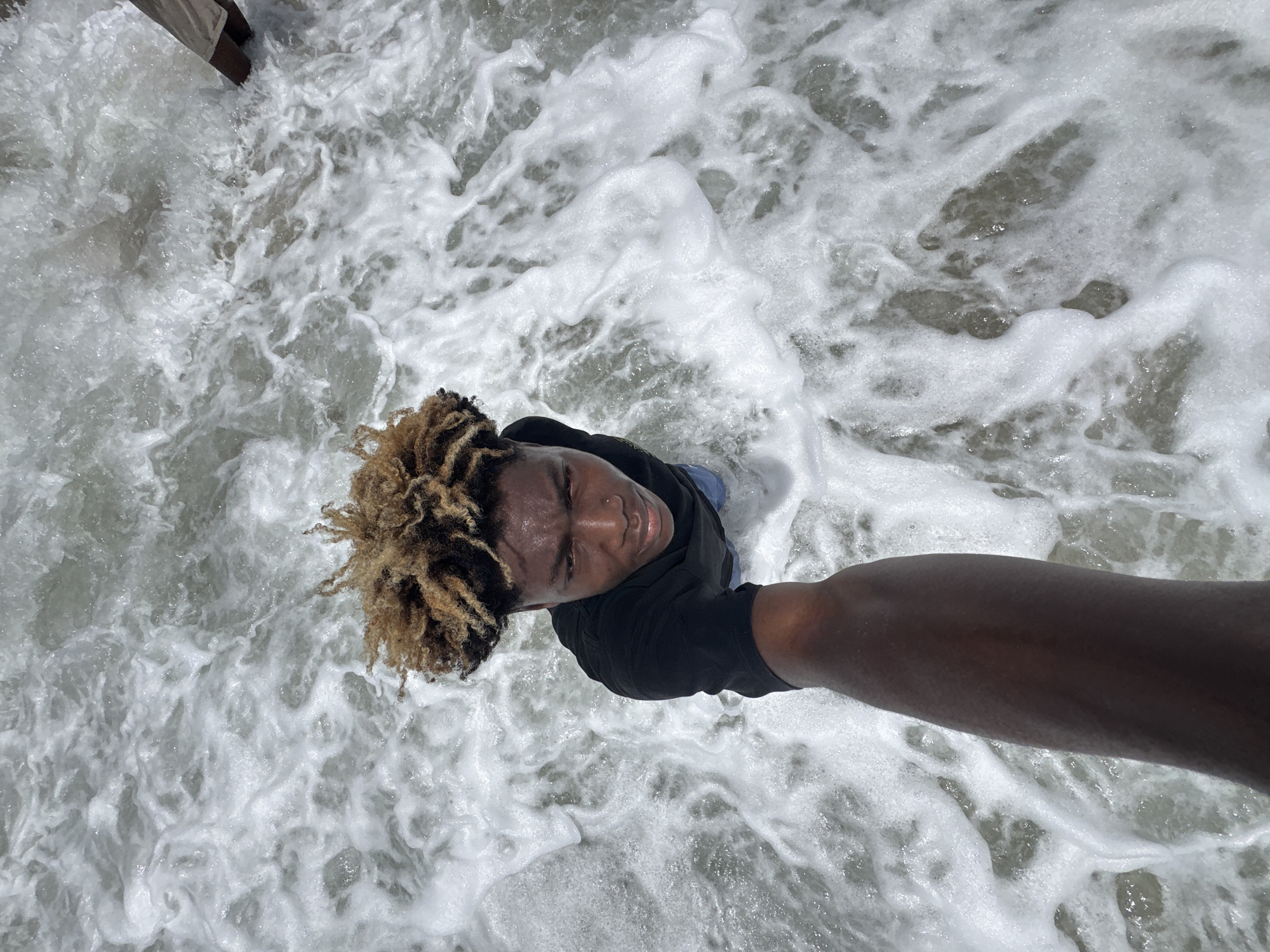 A low-angle, full shot photograph depicts a person standing in foamy, white ocean water. The person is dark-skinned with light brown and dark brown dreadlocks, wearing a dark-colored shirt and light blue pants. The person is facing the camera, taking the picture from a high, downward angle. The background is a sea of white foamy water over a gray ocean floor. The location is assumed to be on a beach, Alaguntan, Nigeria. The sky is not visible in the shot.