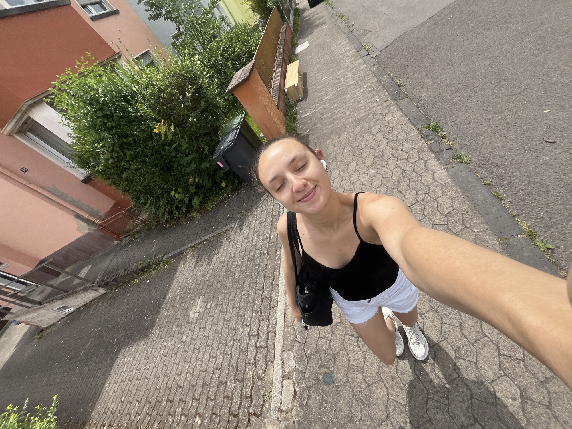 A young woman takes a selfie on a street in Saarbrücken, Germany. She is wearing a black tank top, white shorts, and white sneakers. A black bag is slung over her shoulder. She is smiling with her eyes closed. A paved street and sidewalk are visible. A pinkish-red building and greenery line the left side of the street. A tall, brown fence and a paved asphalt road are on the right. Trash bins and vegetation are visible behind the fence.