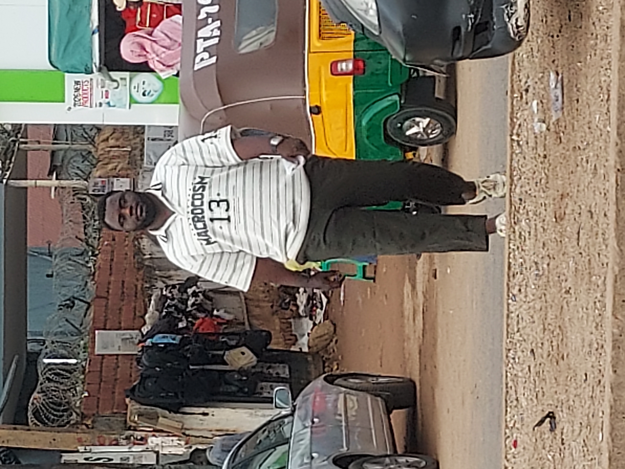 A man in a white and black striped shirt and dark pants stands in front of a parked silver car and a brown, yellow, and green auto rickshaw. The man faces the camera. Behind him, to the left, is a pile of clothing and various objects. The scene appears to be on a street, based on the curb and the presence of vehicles. The auto rickshaw displays "PTA-7" on its side. The location is assumed to be Jos, Nigeria.