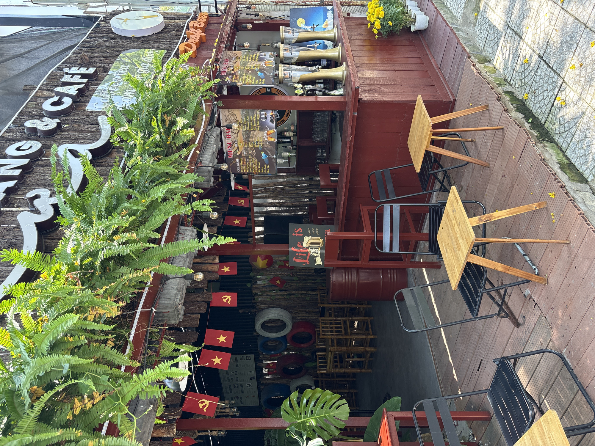 This media file captures an outdoor cafe or bar setup in Đà Nẵng, Vietnam, likely during the daytime. The scene presents a casual, inviting atmosphere with no people visible.
The cafe features a wooden facade with a large sign partially visible, displaying "NG & CAFE," and likely "Cơm gia đình" (family meal) though OCR rendered it as "Com glading." Another prominent sign advertises "Draft Beer." The main structure is made of dark stained wood, with a bar counter equipped with beer taps, bottled beverages, and glasses. Hanging "MENU BAR" signs list cocktails such as "MARGARITA," "Long Island," "Blue Hawaii," "Gordon's Gin," "Bacardi Light," and "Jim Beam." Another sign prominently reads "Life is HAPPY NEW BEER!".
Seating consists of several small square wooden tables paired with black metal folding chairs, arranged on a wooden deck adjacent to a concrete sidewalk. The decor includes lush green ferns and a large monstera leaf, as well as potted yellow flowers, adding to the natural aesthetic. Notably, numerous small red flags with a yellow star (Flag of Vietnam) and yellow hammer and sickle (emblem of the Communist Party of Vietnam) are strung as decorations. Used tires, painted red, blue, and grey, are stacked beneath the main structure, possibly repurposed as decor or seating elements. Bright sunlight illuminates the area, casting shadows, suggesting it is midday or early afternoon.
