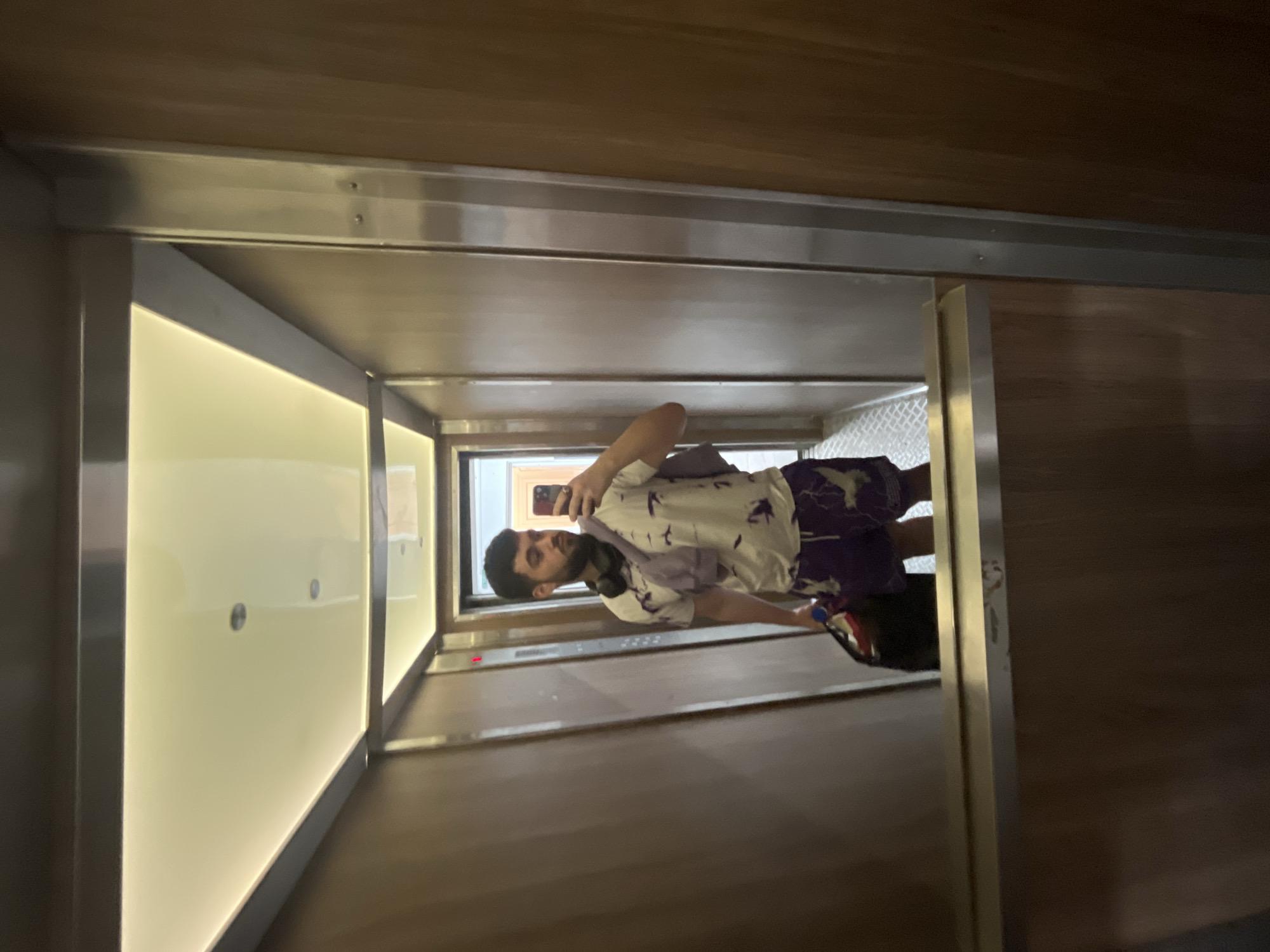 The image depicts a person taking a selfie in an elevator. The person has dark hair and a beard, wearing a patterned shirt and shorts. They are holding a phone up to take the picture. The elevator appears to have wooden walls and a lit-up panel. The precise location in Sofia, Bulgaria, is not discernible from the image.