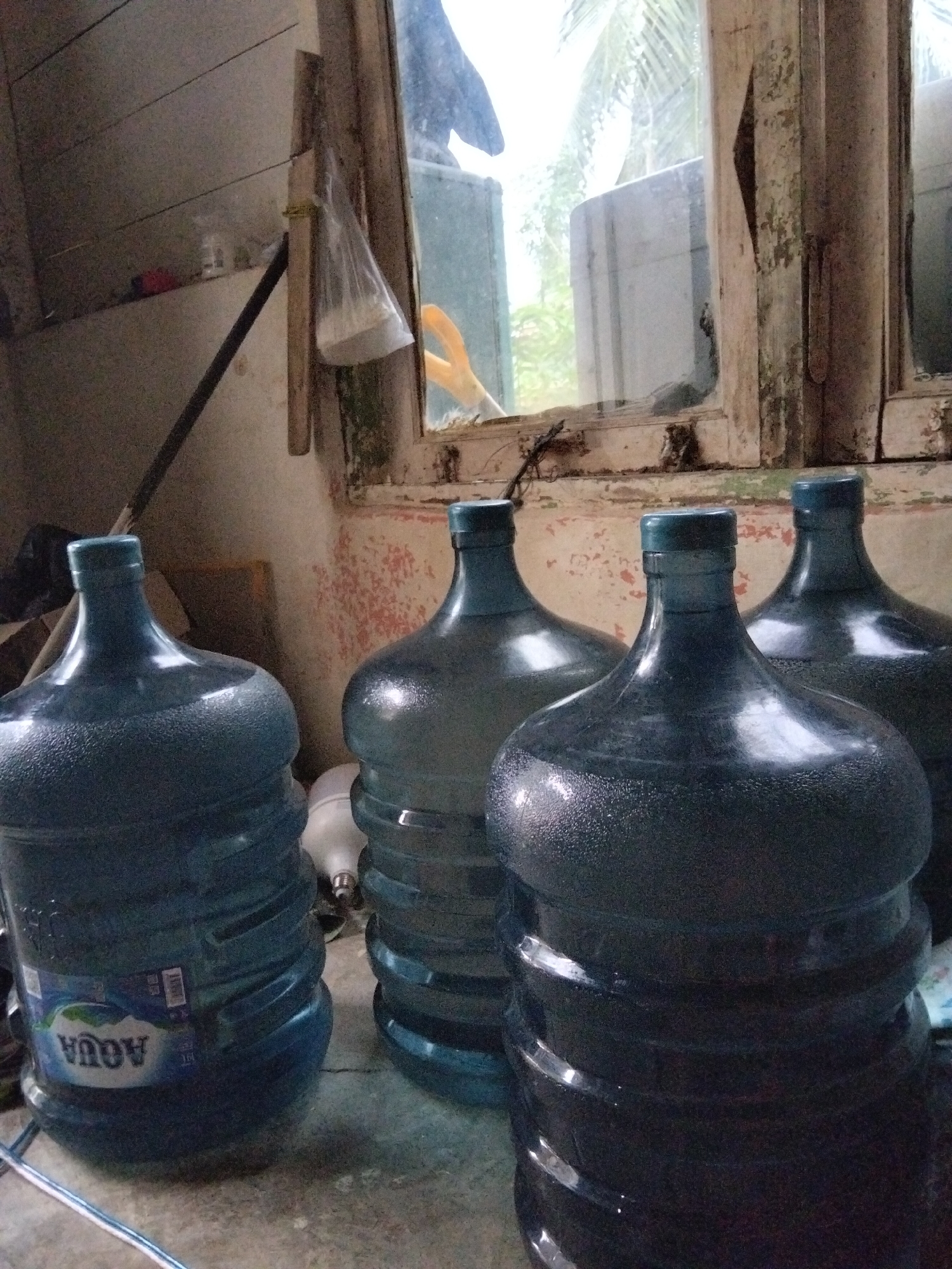 The image shows four large, blue plastic water jugs in what appears to be an interior space. The jugs are standing upright, with their caps visible. The jug on the far left has a label that reads "AQUA" in white lettering against a green and blue background. In the background, a window with dirty panes reveals foliage and trees outside, suggesting a natural or semi-natural setting. The window frame is weathered and peeling. To the left of the jugs, a dark pole leans against a wooden structure. The floor is concrete and appears somewhat dusty. A white object, possibly a lightbulb, is partially visible between the second and third jugs from the left. The overall lighting is dim, with light streaming in from the window. There are no people visible in the image. The scene suggests a storage area or a less-maintained part of a building. The presence of the "AQUA" label indicates a common brand of bottled water in Indonesia. While the image does not explicitly show the city of Benuaraja, Indonesia, the presence of the Aqua brand is consistent with locations in Indonesia.