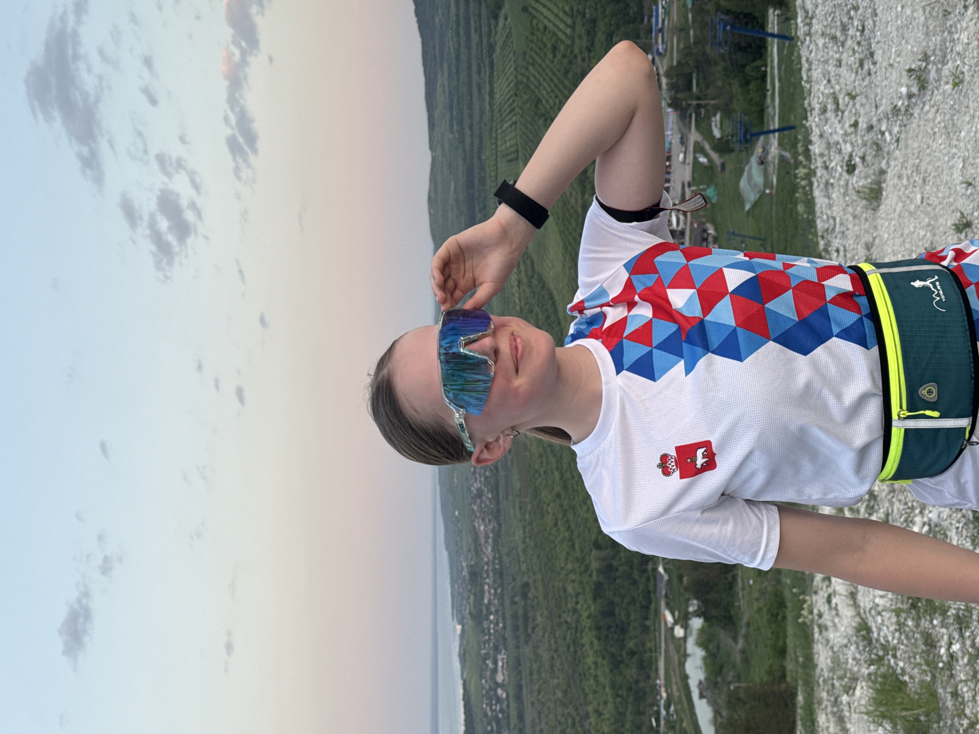 A young woman stands on a rocky precipice overlooking a cityscape, lake, and forested areas. She wears reflective sunglasses, a patterned shirt with a regional symbol on the upper left, a black wristband, and a green hip pack. Her hand is positioned near her sunglasses. The setting is outdoors with the sky visible, suggesting it's evening. The location appears to be in Khvalynsk, Russia.