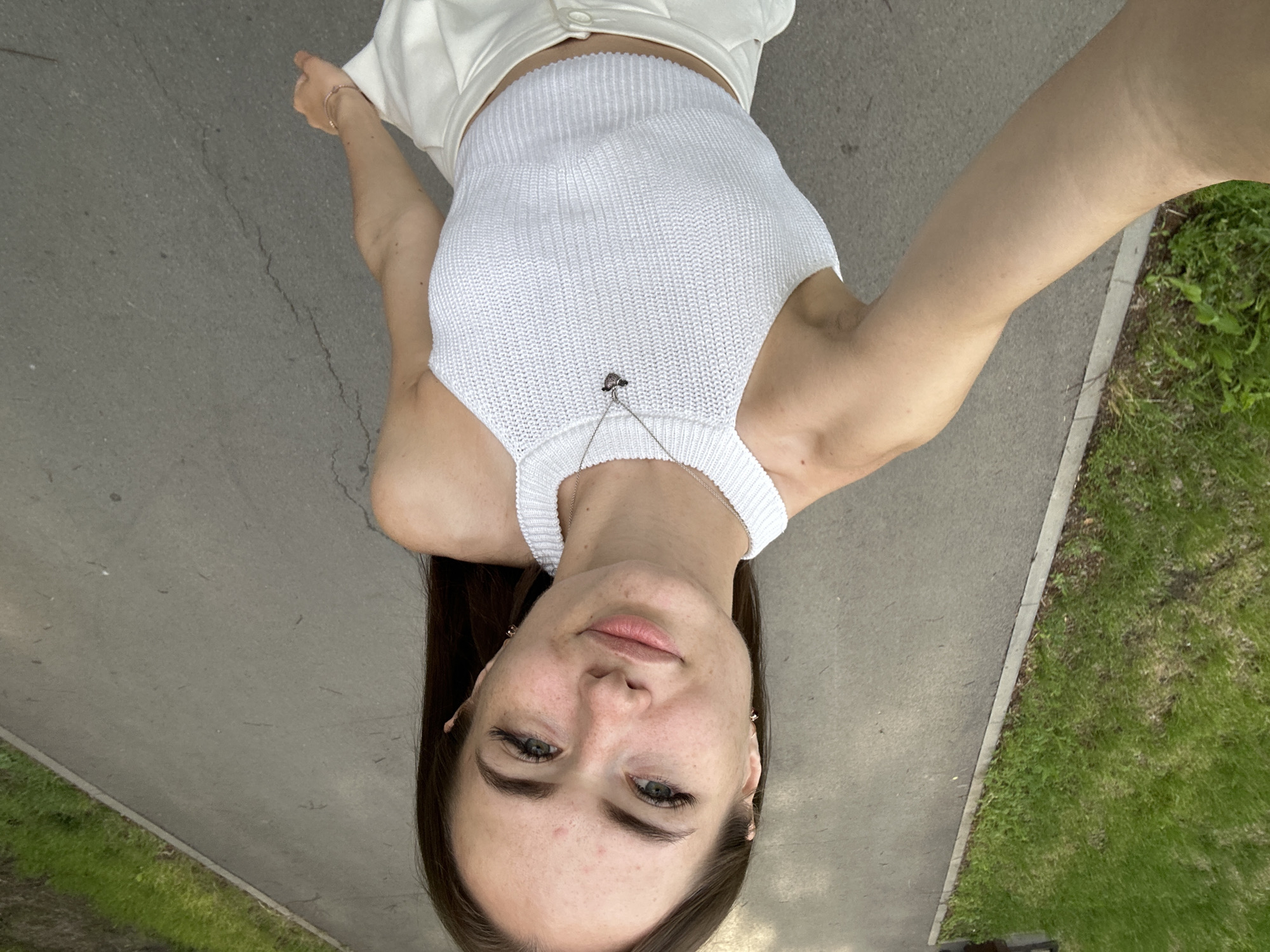 A young woman with long, dark hair is captured from a high angle, taking a selfie as she walks along a paved pathway. She is wearing a white knit halter top and white pants. The pathway is bordered by a strip of green grass on the left side of the frame. The overall scene suggests a casual outdoor setting, possibly a park or a quiet street.