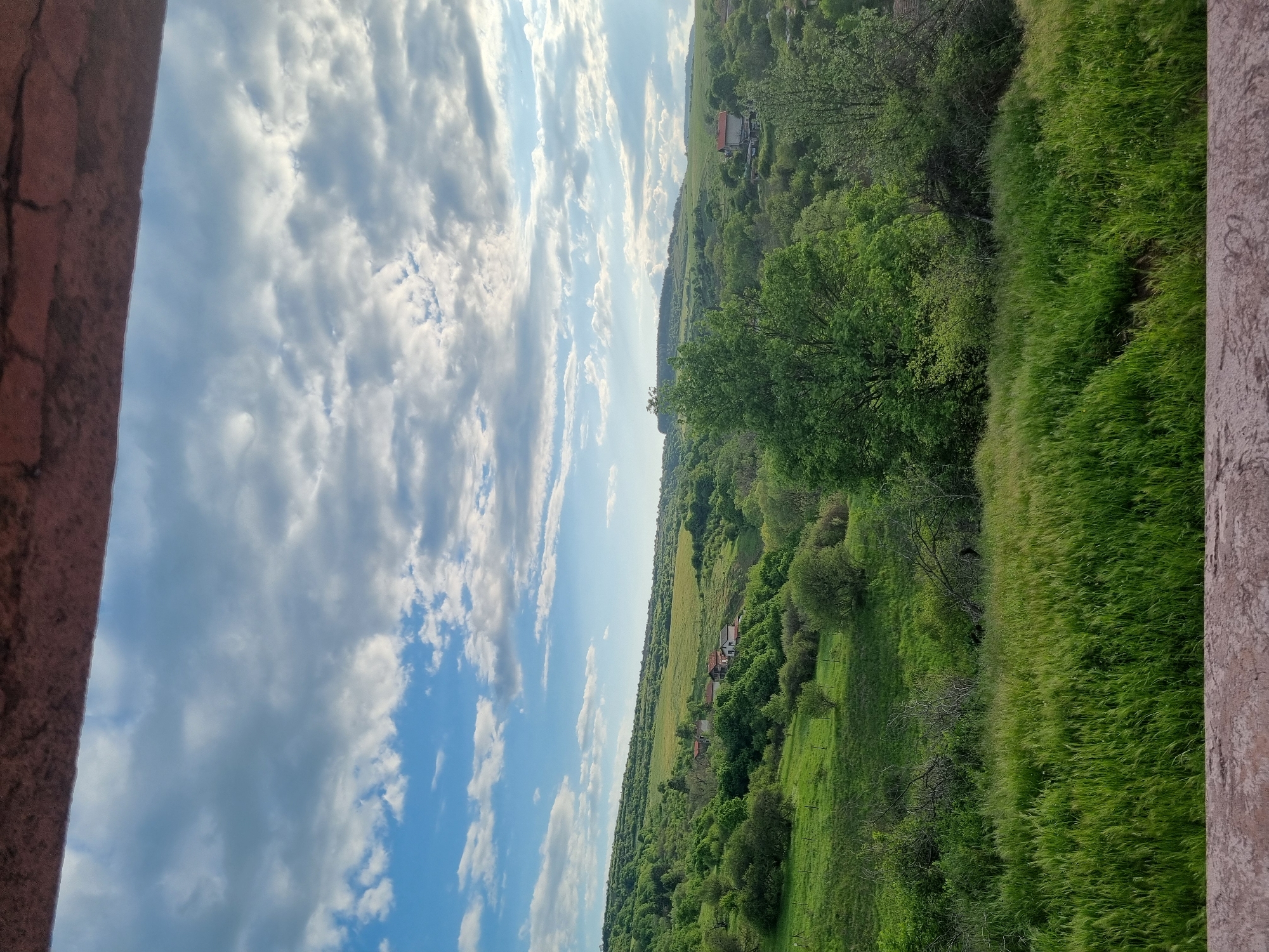 The image shows a landscape view from an elevated position, possibly a building or tower. The foreground features part of a stone structure. The landscape consists of a rolling green terrain with fields, trees, and vegetation. A few buildings are visible in the distance. The sky is blue with scattered clouds. The image is taken in the city of Gurgulyat, Bulgaria.