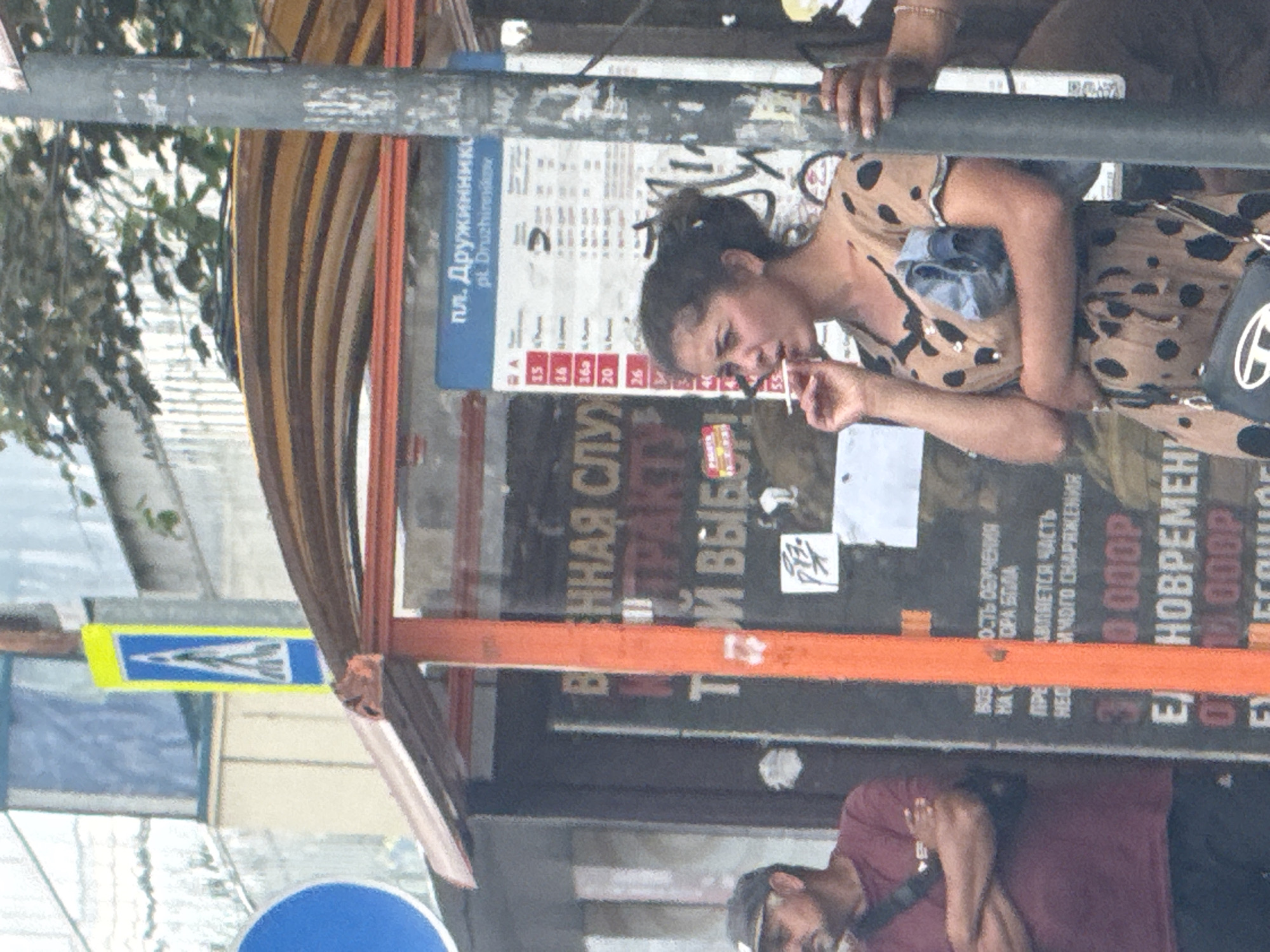 The image depicts a street scene, likely in Rostov-on-Don, Russia, centered around a bus stop shelter. An adult woman with dark hair, wearing a polka-dot dress and holding a cigarette, stands to the right of the frame. To her left, an adult male wearing a burgundy t-shirt and a dark baseball cap is visible, with his arms crossed. Behind the woman, a blue and yellow pedestrian crossing sign is affixed to a pole. The bus stop shelter has an orange-red frame and a brown, curved roof. A bulletin board displaying a schedule and transit information is attached to a gray pole, partially obscuring the woman. The background shows building facades, some with striped patterns, and a tree with green foliage overhead. Graffiti is visible on the gray pole. The overall impression is a typical urban environment.