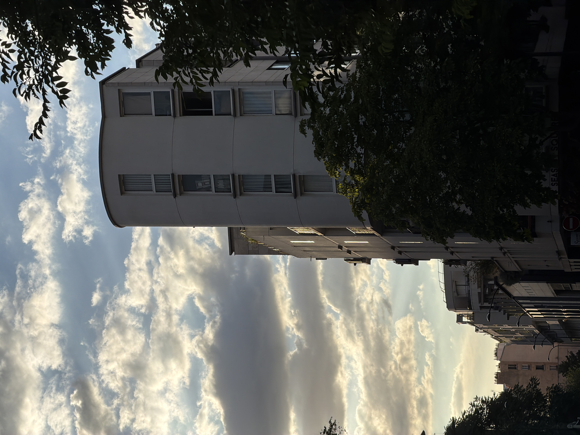This is a medium shot, taken from street level, of buildings and trees in Paris, France. The sun is setting, casting a warm glow on the clouds and the upper portions of the buildings. The sky is filled with dramatic clouds, with streaks of sunlight breaking through.
In the foreground, on the right side of the image, a large tree with dark green leaves obscures part of a modern white building with rounded corners and multiple stories. This building has several rectangular windows, some with blinds, and some reflecting the sky. To the left of this building, the top of a dark green tree can be seen, partially silhouetted against the sky.
Further down the street, a row of buildings of varying heights and architectural styles are visible. Some have balconies and awnings. The street is lined with streetlights. The building on the far left of this row is lighter in color and appears to have a more traditional Parisian style with several windows. Further down the street, the buildings become darker and more modern. One building has a sign that reads "SEASON SQUARE".
The overall atmosphere is serene and urban, capturing a moment of transition between day and night in a Parisian cityscape. There are no people visible in the image.