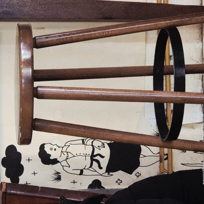 The image displays an indoor scene located in Strasbourg, France. A prominent, dark brown wooden bar stool occupies the central and right portions of the frame. It features a round, polished wooden seat supported by four slender, tapered wooden legs. A black metal ring serves as a footrest, encircling the legs approximately halfway down.
In the background, a light-colored wall, possibly off-white or cream, exhibits a black-and-white mural. The mural depicts a line drawing of a woman in profile, facing towards the right. She has short, dark hair and wears a collared, short-sleeved shirt with a dark sash or apron tied around her waist, and a dark skirt. Scattered around the woman are additional graphic elements, including stylized black clouds, small crosses, and rudimentary grass shapes. The lower section of the wall shows signs of wear, with visible flaking or textural imperfections in the paint.
To the far left, a dark, possibly black, garment is partially visible, hanging next to a vertical dark brown wooden structure. A similar vertical dark brown wooden element is present on the far right edge of the frame.