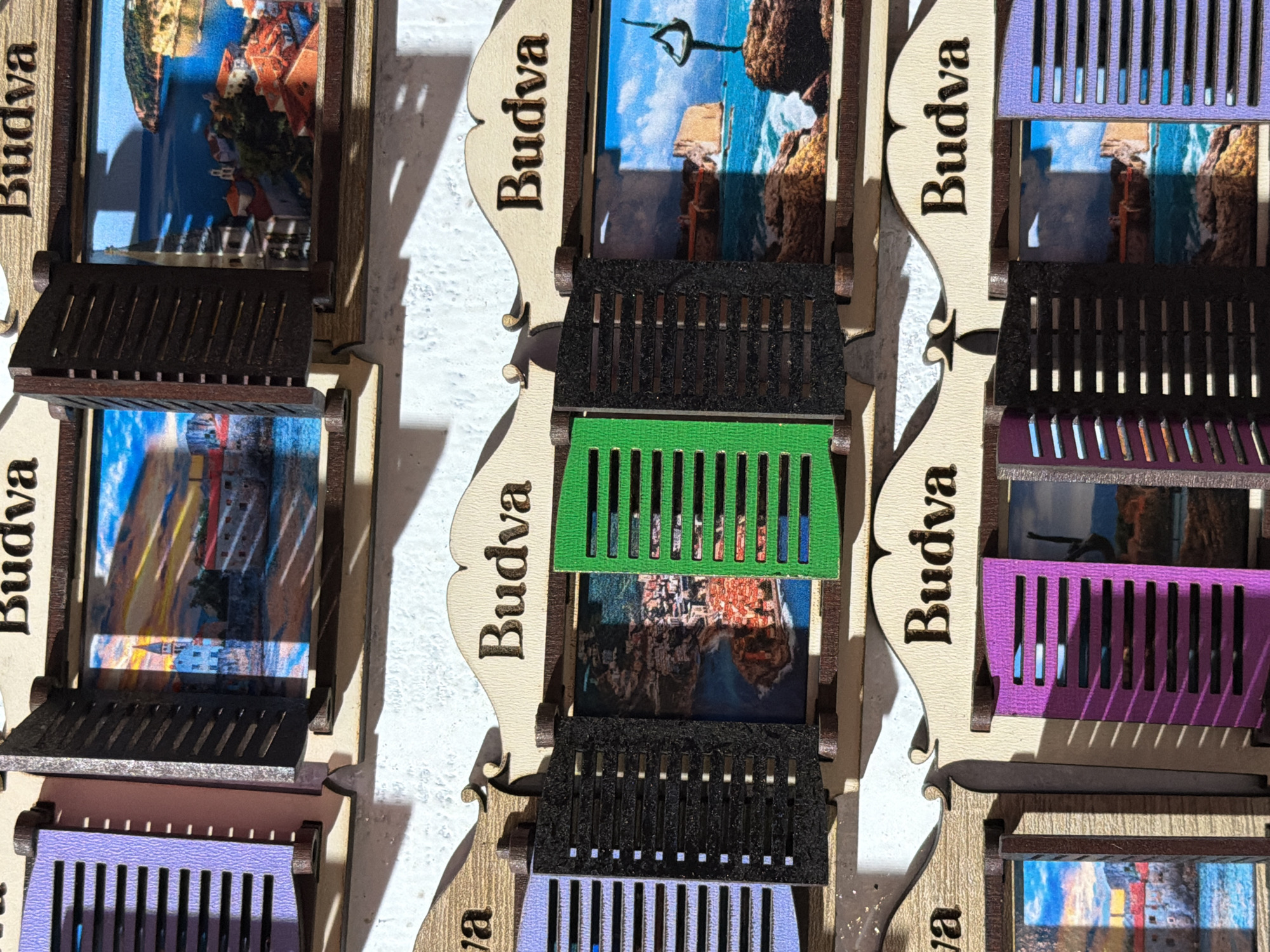 The image displays a close-up view of decorative wooden magnets designed to resemble windows with shutters. Each magnet features a picture of the city of Budva, Montenegro, visible through the open shutters. The word "Budva" is inscribed above each window. The magnets are arranged in multiple rows, suggesting a display for sale. The scene is lit by natural light, casting shadows across the surface.