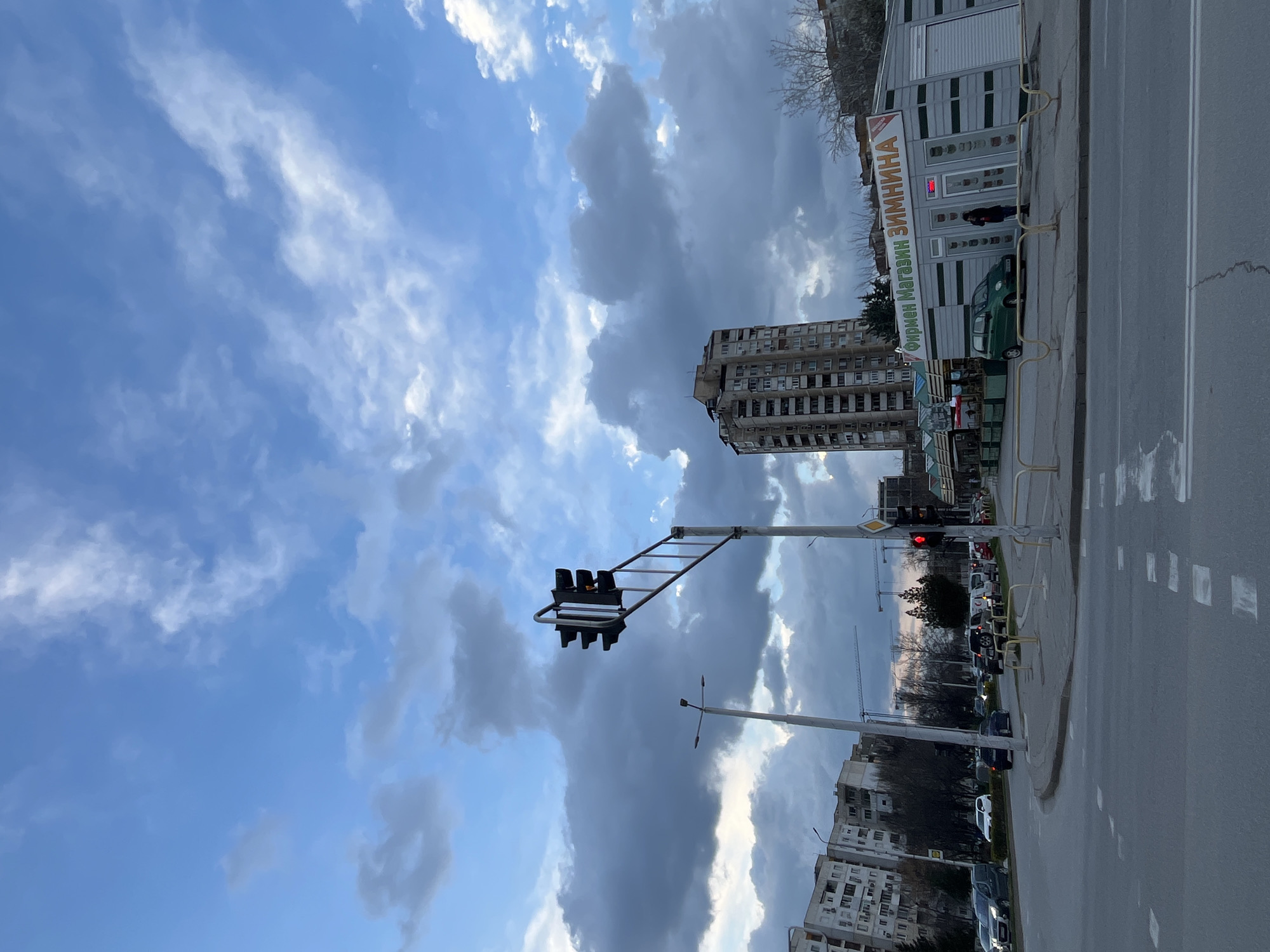 The image displays an urban street scene at the intersection of Tsar Boris III Obedinitel Boulevard and Vasil Aprilov Boulevard in Plovdiv, Bulgaria. The sky is partially overcast with prominent dark gray clouds and interspersed patches of lighter blue.
In the foreground, an asphalt road with visible white dashed and solid lane markings, including pedestrian crossing lines, occupies the lower third of the frame. An overhead traffic light structure, supported by a pole on the right side of the road, shows a red light illuminated, indicating a stop for vehicular traffic in the immediate foreground.
To the right, a single-story commercial kiosk with a corrugated gray and white facade is prominent. Its front features the Cyrillic text "ФИРМЕН МАГАЗИН ЗИМНИНА" (BRANDED SHOP ZIMNINA) in red and orange lettering. A small, two-door, dark green vehicle is parked directly in front of this kiosk. One individual, dressed in dark clothing, stands facing the shop's facade. Yellow metal barriers line the edge of the sidewalk in front of the kiosk.
Behind the kiosk and slightly to the left, a tall, multi-story residential building with numerous windows and balconies extends upwards. On the left side of the intersection, a row of multi-story, light-colored residential apartment buildings lines the street. Bare deciduous trees, suggesting winter or late autumn, are present along the median and sidewalks. Multiple vehicles, including cars and vans, are visible on the roads; some are stationary at the red light on the right, while others are on the left-hand street. Streetlights are also visible.