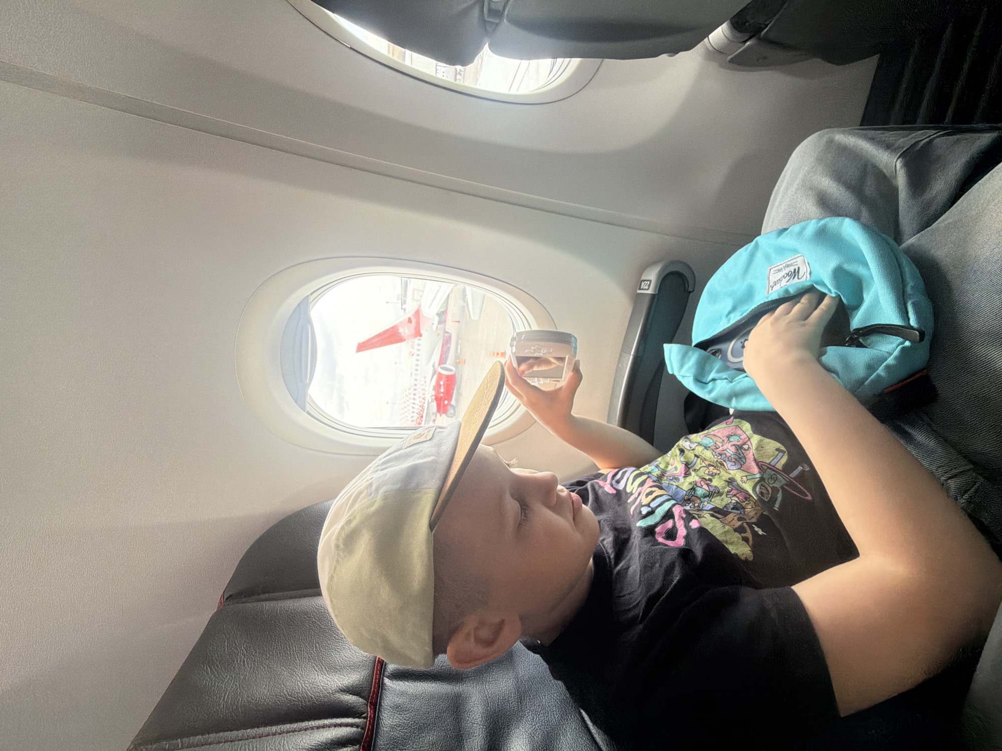 A child is depicted inside an airplane cabin, seated or reclining in a window seat during what appears to be bright daylight. The child, likely a boy, has short light-colored hair and wears a two-toned baseball cap (beige brim, light-colored top), a black t-shirt with a colorful cartoon graphic, and grey trousers. He holds a small, clear container with liquid in his right hand, looking out the oval aircraft window.
Through the window, parts of an aircraft's red and white tail fin or fuselage and some ground equipment are visible, suggesting the plane is stationary at an airport. The cabin interior is light-colored and appears clean.
Resting on the child's lap or beside him is a light blue backpack or bag, on which the brand name "Herschel" is clearly visible. One of the child's hands is reaching inside this bag. A white label with the alphanumeric "V22" is affixed to the side panel next to the window. The child's expression is neutral, possibly indicating curiosity or relaxation as he observes the outside world. The provided context indicates the location is Saint Petersburg, Russia.