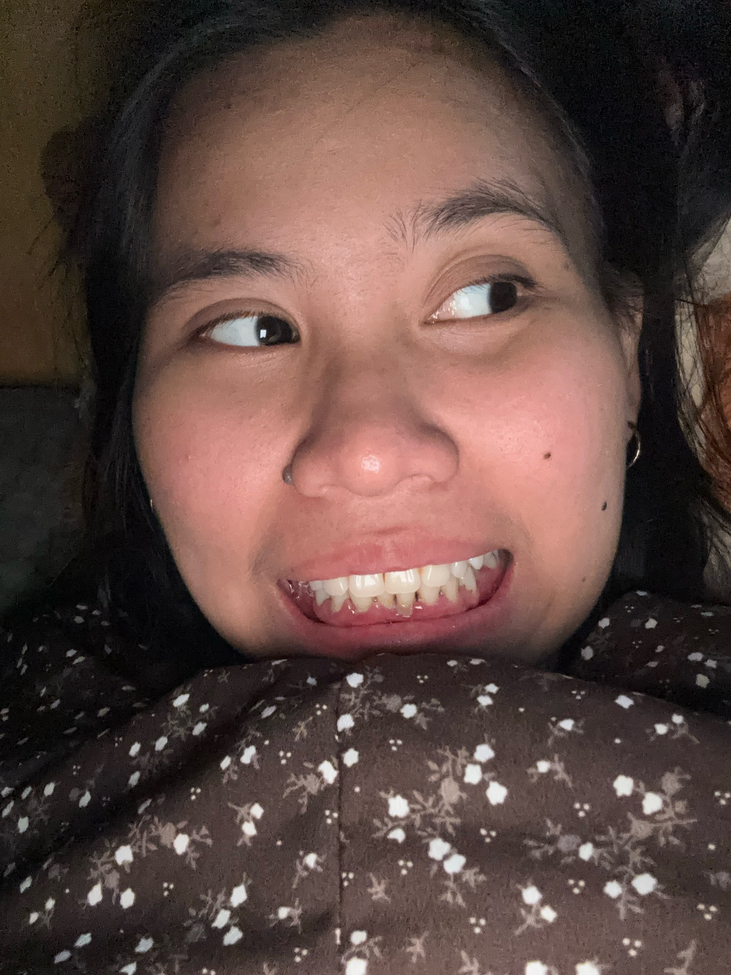 The image is a close-up selfie of a young woman lying down, with her head tilted to the side. She is looking to her right with her eyes wide. She has dark hair that is partially visible, and she is wearing a dark brown top with a white floral pattern. Her mouth is slightly open, revealing her teeth. The lighting is dim, suggesting the photo was taken indoors, possibly in a bedroom. The background is dark and out of focus, with a hint of what appears to be a blanket or bedding. There are no discernible landmarks or elements that would indicate a specific location in East Jakarta, Indonesia.