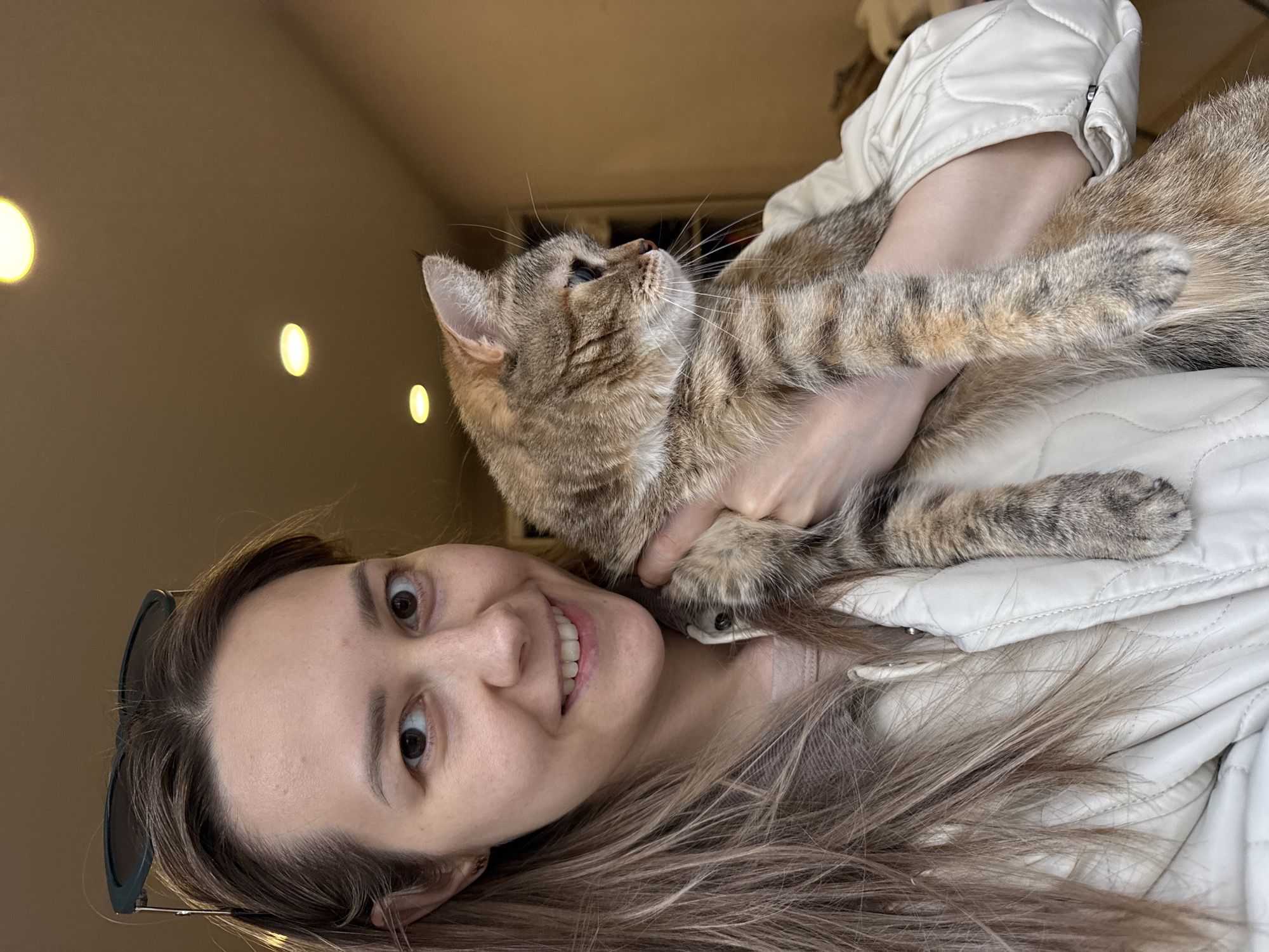 A young woman holds a tabby cat in an indoor setting, captured in a selfie or casual portrait. The woman, positioned on the right, smiles widely and looks directly at the camera. She has long, light brown hair and dark sunglasses perched on her head. She wears a light-colored, possibly white or off-white, quilted jacket over a light top, with her left hand visible supporting the cat.
The tabby cat, with light brown and grey striped fur, is held close to the woman's chest on the left. Its head is turned upwards and to its right, away from the camera, displaying prominent whiskers and a curious gaze. Its front paws rest on the woman's arm or jacket.
The background consists of a plain, warm brown or beige wall or ceiling, indicating an indoor location. Several blurred, circular yellow light sources are visible in the upper background. The woman appears happy and content, while the cat seems relaxed. The image does not contain any discernible text or specific outdoor location cues, though the context provided is Chaikovsky, Russia.