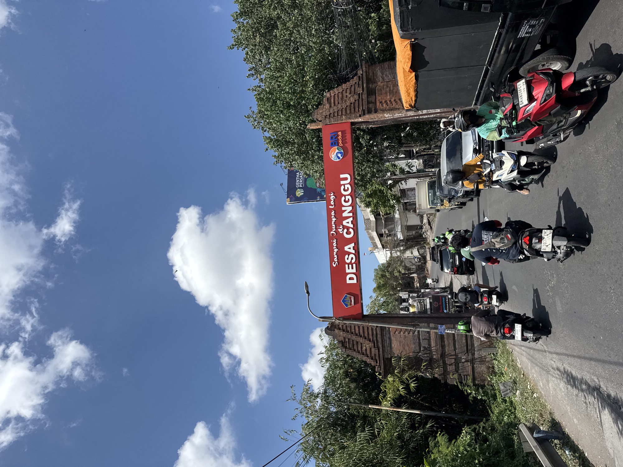 The image depicts a street scene in North Kuta, Indonesia, under a bright blue sky with scattered white clouds. A prominent red archway, inscribed with "Sampai Jumpa Lagi di DESA CANGGU" and the BRI logo, spans the road, signifying an entrance. Several motorcycles are visible, with riders wearing helmets, navigating the street. Some motorcycles are moving, while others are stationary in traffic. On the right side of the road, a large truck with an orange tarp is parked. Lush green trees and vegetation line the sides of the road, and in the background, several buildings and a billboard are visible. The scene appears to be a typical day on a busy street in a tropical location.