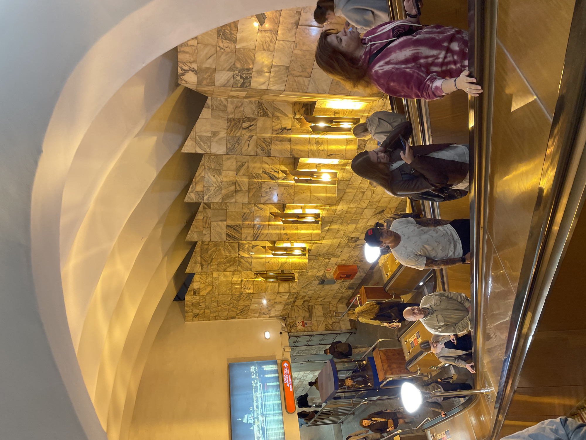 The image captures an indoor scene of an escalator within what appears to be a metro station. A diverse group of individuals is visible on the escalator, some riding up and others down. The walls of the station are adorned with a patterned tile design, likely marble, creating a geometric and visually striking aesthetic. Lighting fixtures are integrated into the wall design, adding to the ambiance. A digital display screen is present, providing information, possibly regarding train schedules or station navigation. The overall environment suggests a busy urban setting with people utilizing public transportation. The location is Unknown, Unknown.
