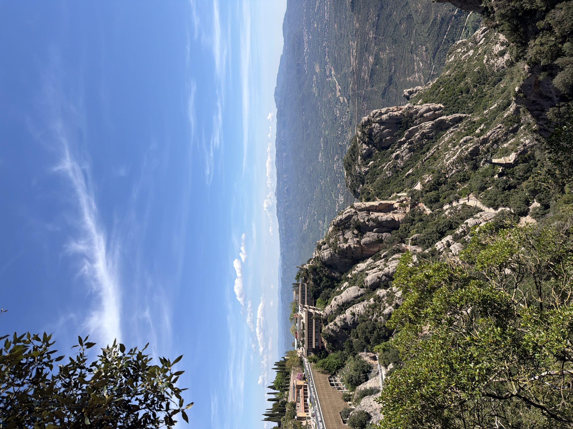 This image captures a panoramic view of the Montserrat mountain range in Collbató, Spain, under a bright, clear blue sky with sparse, wispy cirrus clouds. The scene is observed from a high vantage point, looking out over a dramatic landscape characterized by distinctive, jagged rock formations covered with dense green Mediterranean vegetation.
In the midground, several modern buildings with multi-level terraces and flat roofs are integrated into the steep, rocky slopes, suggesting a monastery complex or visitor facilities. Winding paths or roads are visible along the mountainside, connecting these structures. The foreground features more of the characteristic rocky terrain and lush green trees, with some foliage partially framing the bottom left and right of the view. In the far distance, a hazy horizon line separates the sky from further undulating mountain ranges and valleys.
The bright sunlight suggests midday or early afternoon, casting distinct shadows and highlighting the rugged textures of the rocks. The weather is clear and sunny. There are no people visible in the image, nor is any discernible text present. The overall impression is one of grandeur, natural beauty, and the serene integration of human structures within a powerful natural environment.