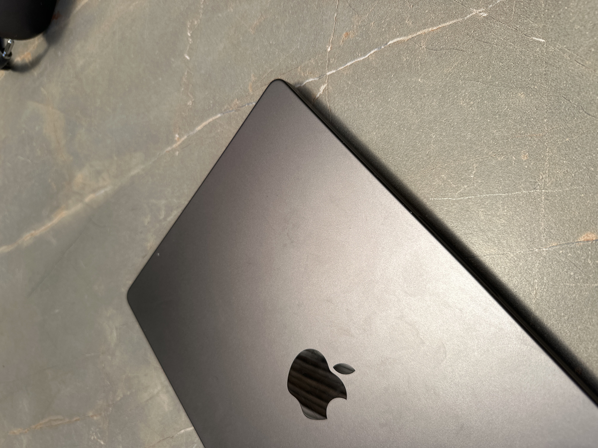 A top-down, close-up shot reveals a closed, dark gray laptop, likely a MacBook, positioned on a gray, stone-like surface. The Apple logo is prominently displayed on the laptop's lid. Part of a black object, possibly headphones, is visible in the upper left corner. The setting appears to be indoors, possibly a workspace or home. The location is unknown, not related to the city of Bnay Zion, Israel.