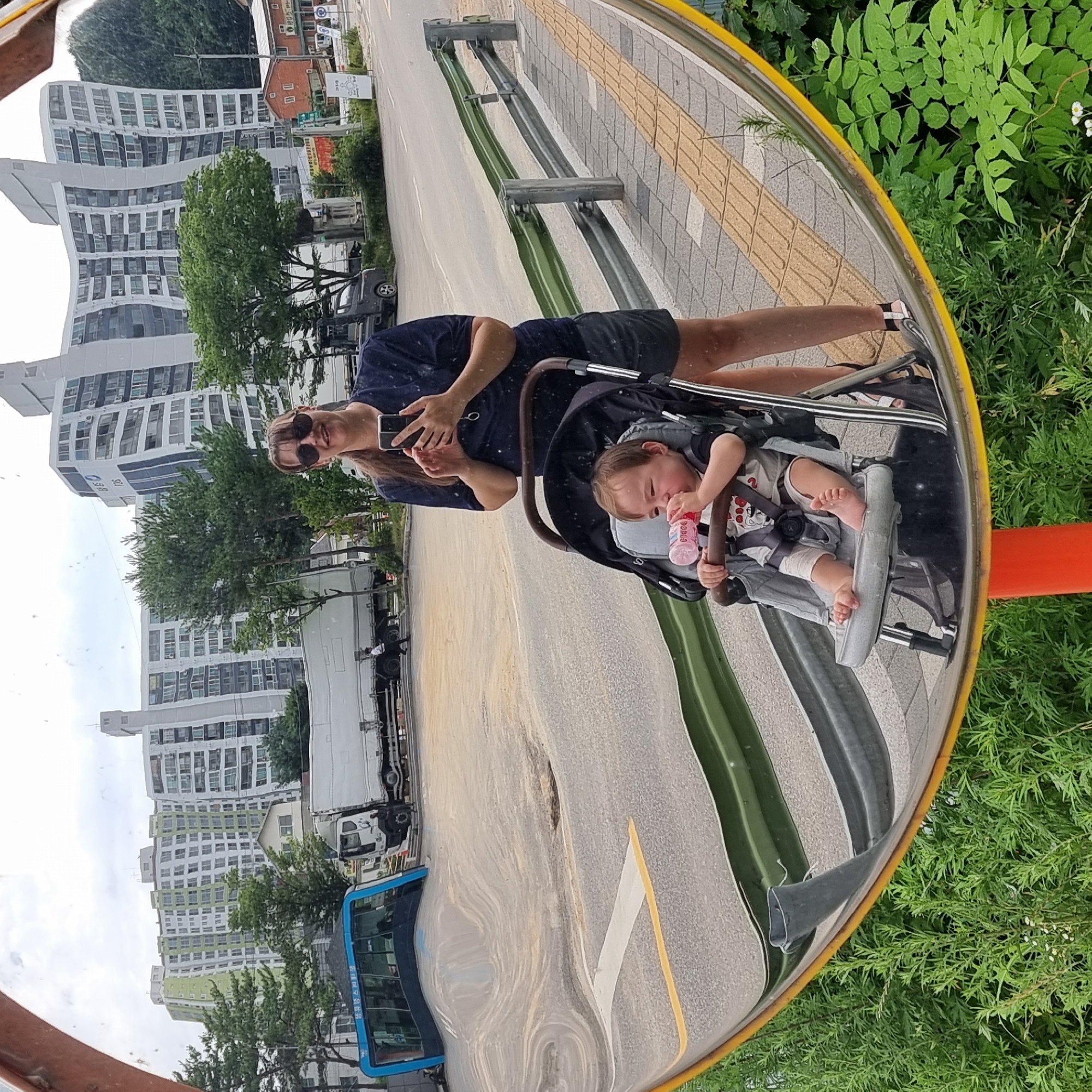 This is a wide-angle, distorted reflection in a convex mirror, likely a traffic mirror. The reflection shows a woman with sunglasses and dark hair in a ponytail holding a smartphone and taking a selfie. She is wearing a dark blue t-shirt and dark shorts. Next to her, in a stroller, is a young child, likely a toddler, drinking from a pink bottle. The child is wearing a light-colored t-shirt with a graphic print and has bandages on their knees. The background of the reflection shows a street scene with apartment buildings, trees, a bus, and a truck. The buildings in the background appear warped and wavy due to the mirror's curvature. The foreground shows green foliage and the bottom edge of the convex mirror supported by an orange pole. The scene is set in Dunnae-myeon, South Korea, as indicated by the text on the bus.