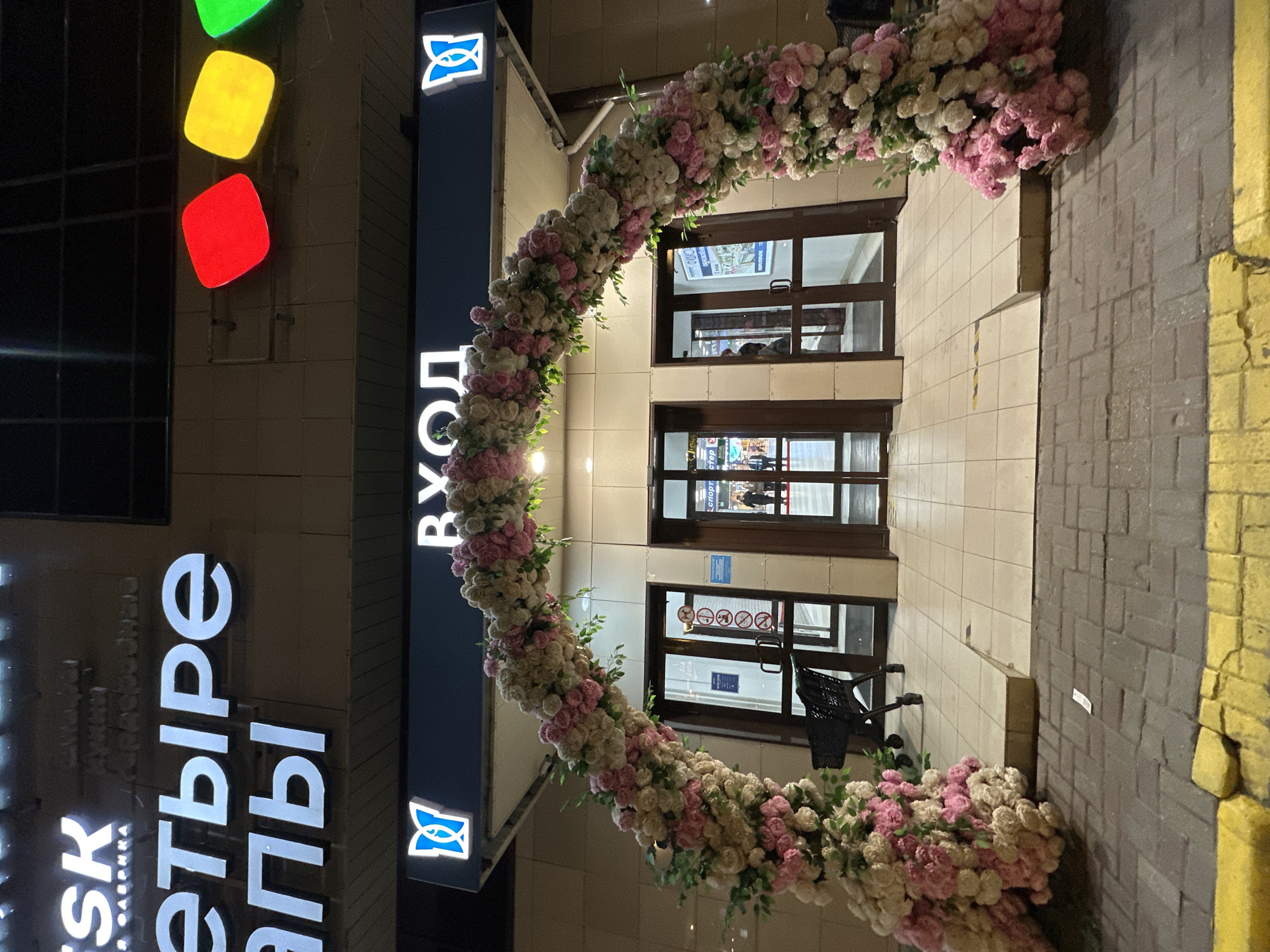 The image shows the entrance to a store, possibly in Moscow, Russia. The entrance is decorated with a large arch of pink and white flowers. A sign next to the entrance reads "ВХОД," which means "Entrance" in Russian. A sign for the store reads "Четыре Лапы", a chain of pet stores in Russia. The entrance has three sets of doors, allowing a view into the store. There are traffic-light shaped lights on the left, with red, yellow, and green sections.