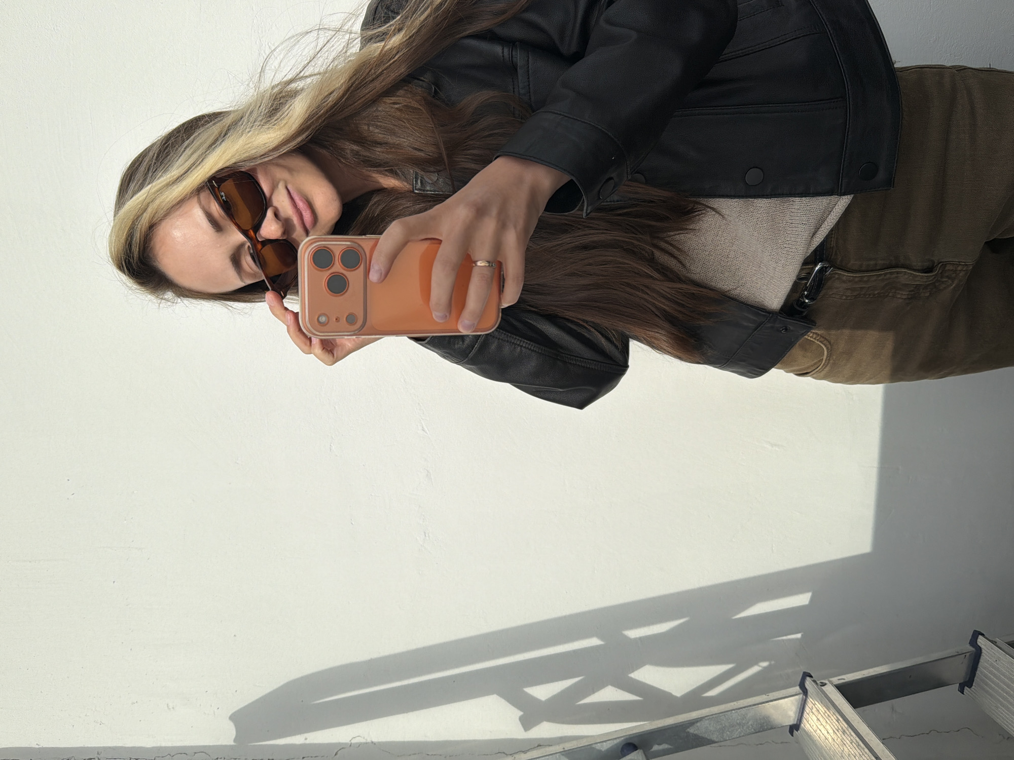 This is a vertical, medium shot of a young woman taking a selfie in a brightly lit, indoor setting. She is positioned in the upper right quadrant of the frame, looking down at her phone. The woman has long, wavy blonde and brown hair that falls over her shoulders and frames her face. She is wearing oversized, brown-tinted sunglasses, a light beige sweater, and a black leather jacket. On her left hand, she wears a thin, gold ring. Her phone, held in her right hand, has an orange case.
The background is a plain, white wall, casting a shadow from what appears to be a ladder or shelving unit in the lower right corner of the image. The lighting suggests it is daytime, with a strong light source creating distinct shadows. The overall mood is casual and relaxed, indicated by the selfie pose and her relaxed expression. There is no visible text in the image. The context provided is Khimki, Russia, but this is not discernible from the visual information alone.