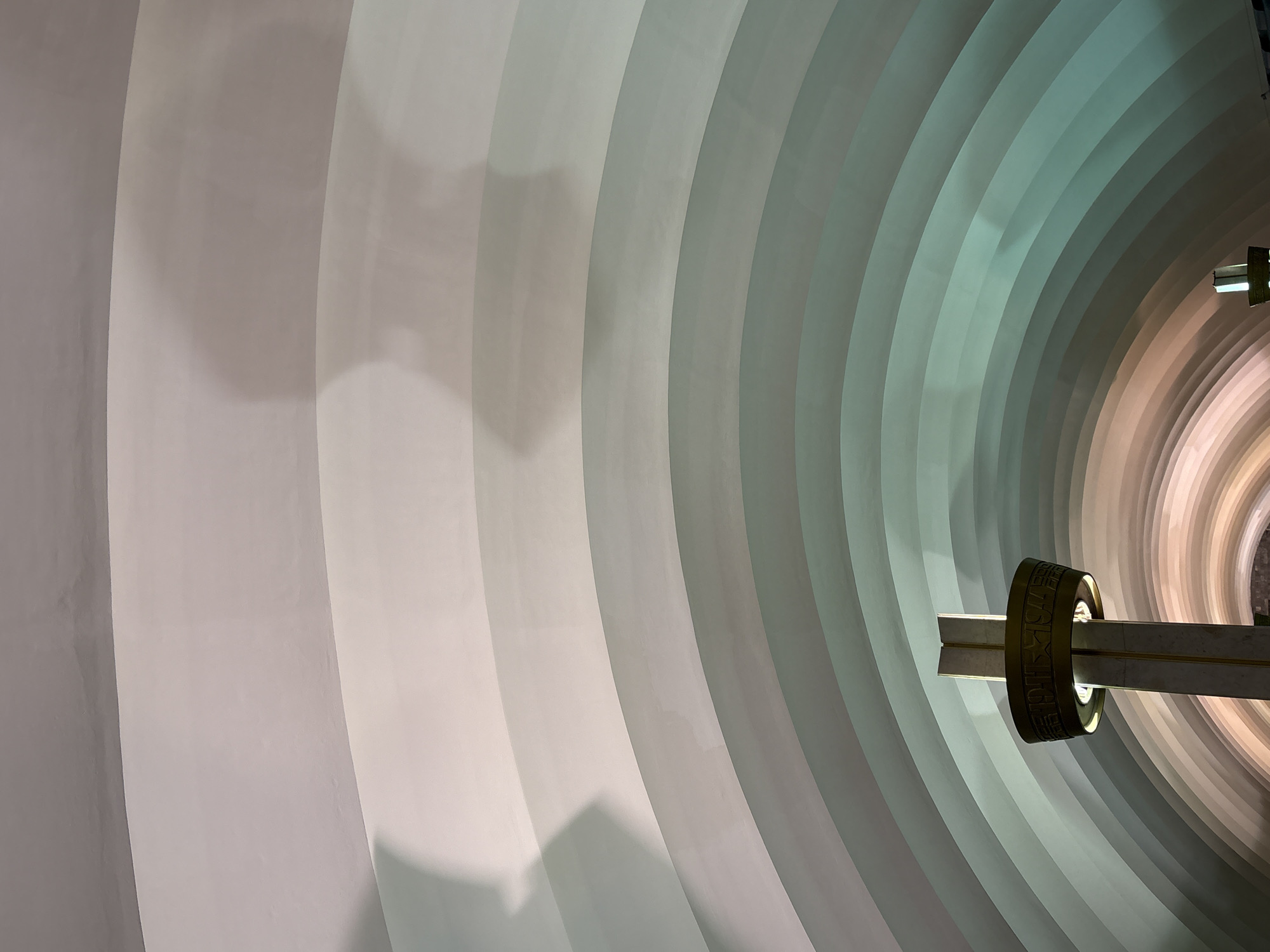 The image depicts the interior of a curved, ribbed architectural structure, likely a tunnel or an underpass, in Saint Petersburg, Russia. The ribbed ceiling creates a series of arching bands that recede into the distance. The lighting creates a gradient of colors, transitioning from warm pinks and oranges at the bottom to cooler blues and greens further up, with soft shadows cast across the ceiling. In the foreground, a portion of a decorative column with a metallic, ring-shaped fixture is visible on the left side. The overall impression is one of modern, possibly monumental architecture, with an emphasis on form and light.