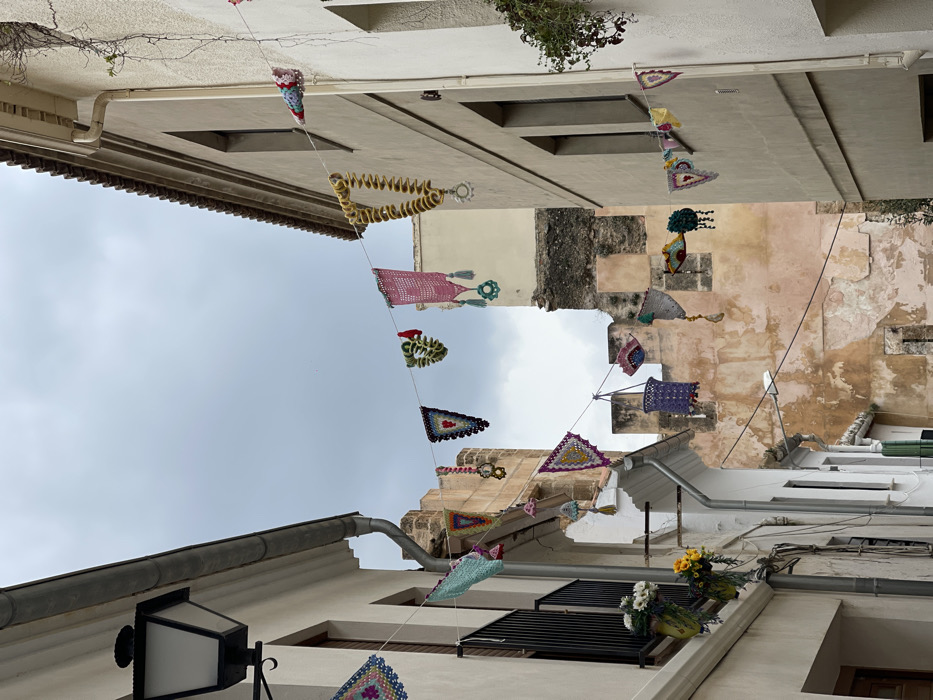 The image displays a narrow street or alleyway in Buñol, Spain, flanked by multi-story buildings under an overcast sky. The street is festively decorated with numerous multi-colored crocheted items strung between the buildings at various heights. These decorations consist of diverse shapes including triangular flags, squares, circular motifs, bell-shaped elements, and a distinctive long, coiled yellow piece, all exhibiting intricate patterns and vibrant colors such as pink, teal, purple, yellow, and green.
On the left side, a light-colored building features a black, ornate street lamp mounted high on the wall, a metallic rain gutter and downspout system, and a window with a black metal balcony railing. Two green hanging containers beneath the balcony hold artificial white and yellow flowers.
The building on the right, also light-colored with a slightly darker tiled roof edge, has a vertical downspout and a vine growing down its wall. Windows with dark frames are visible on both structures.
In the background, at the terminus of the street, a weathered, light reddish-brown stone wall, indicative of an ancient fortification or castle, rises prominently. This structure presents sections of darker stone and visible crenellations. The sky is uniformly grey and cloudy, providing diffuse light to the scene. No individuals are present in the depicted area.