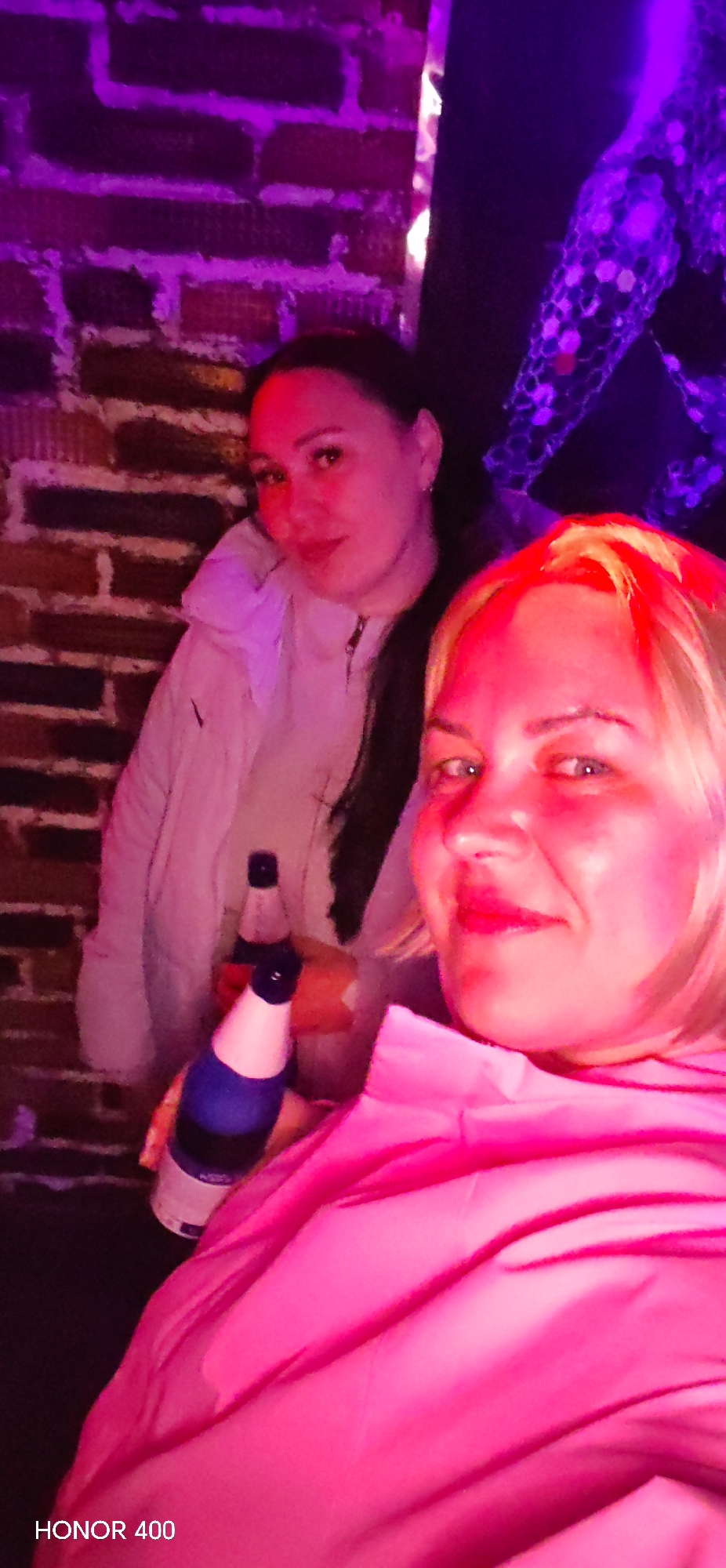 Two women are indoors with a brick wall behind them. The woman in the foreground is wearing a pink jacket and has blonde hair. She is holding a blue and white bottle. The second woman is wearing a white jacket and has dark hair. She is also holding a similar bottle. The scene is lit with a purple hue, particularly noticeable on the brick wall and a decorative element in the background. The location is unknown.