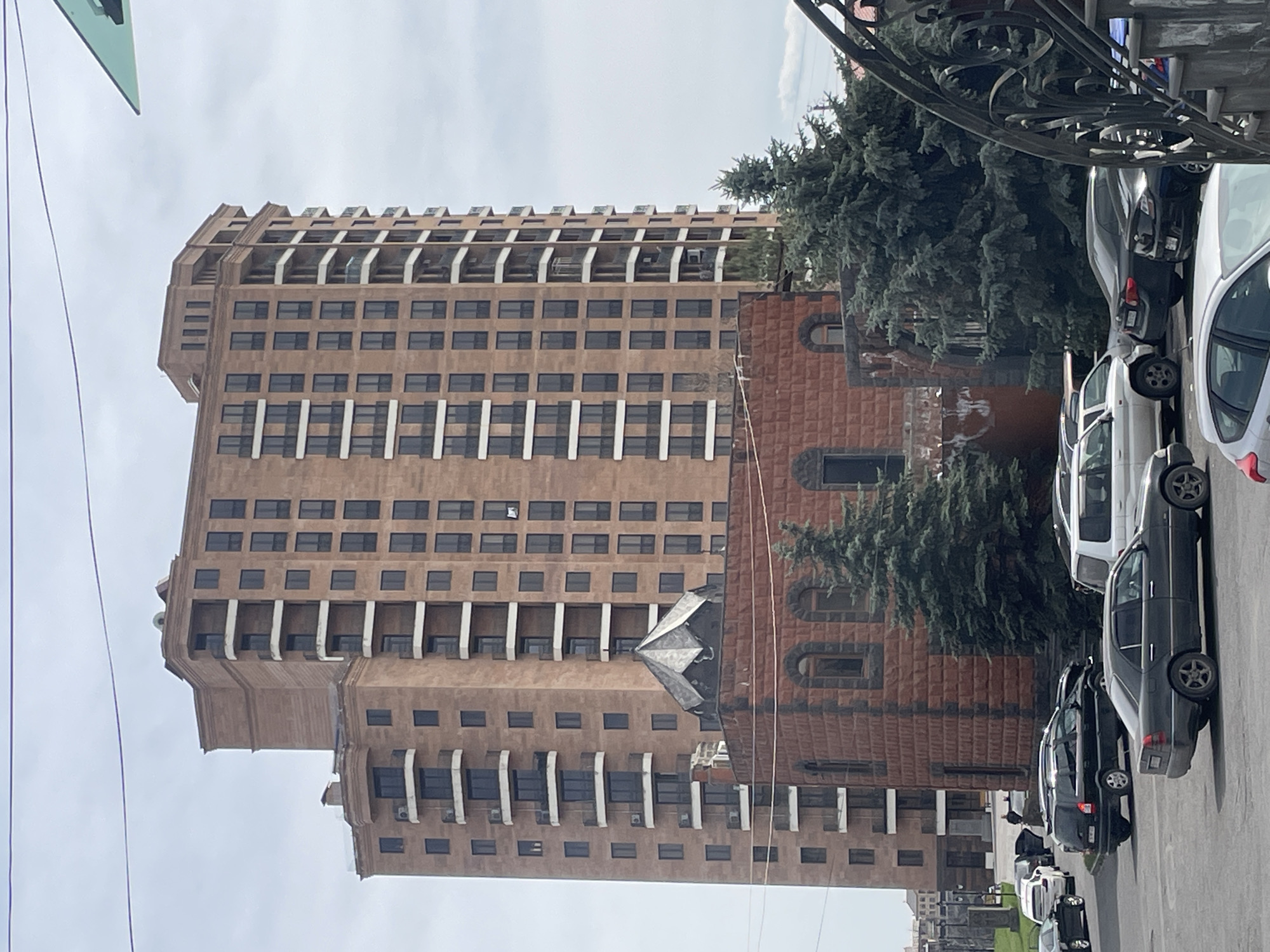 The image shows a tall, brown, multi-story building in Yerevan, Armenia. The building has many windows and white balconies. To the right of the tall building is a smaller building made of red brick. Several trees are adjacent to these buildings. A row of parked cars lines the right side of the image. The sky is overcast.