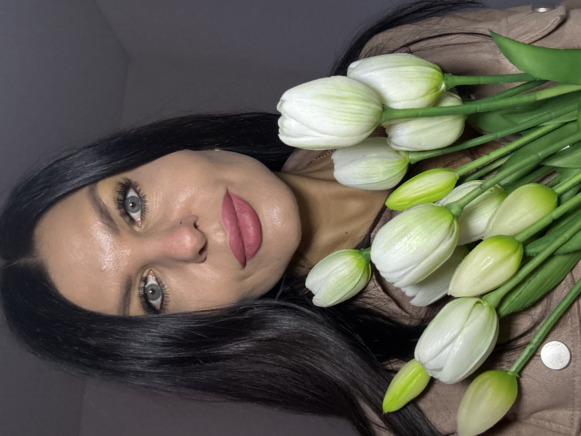 A light-skinned woman with long, dark hair lies on her side holding a bouquet of white tulips with green stems. She has light-colored eyes and wears pink lipstick. She appears to be wearing a tan jacket. The location is not evident from the image.