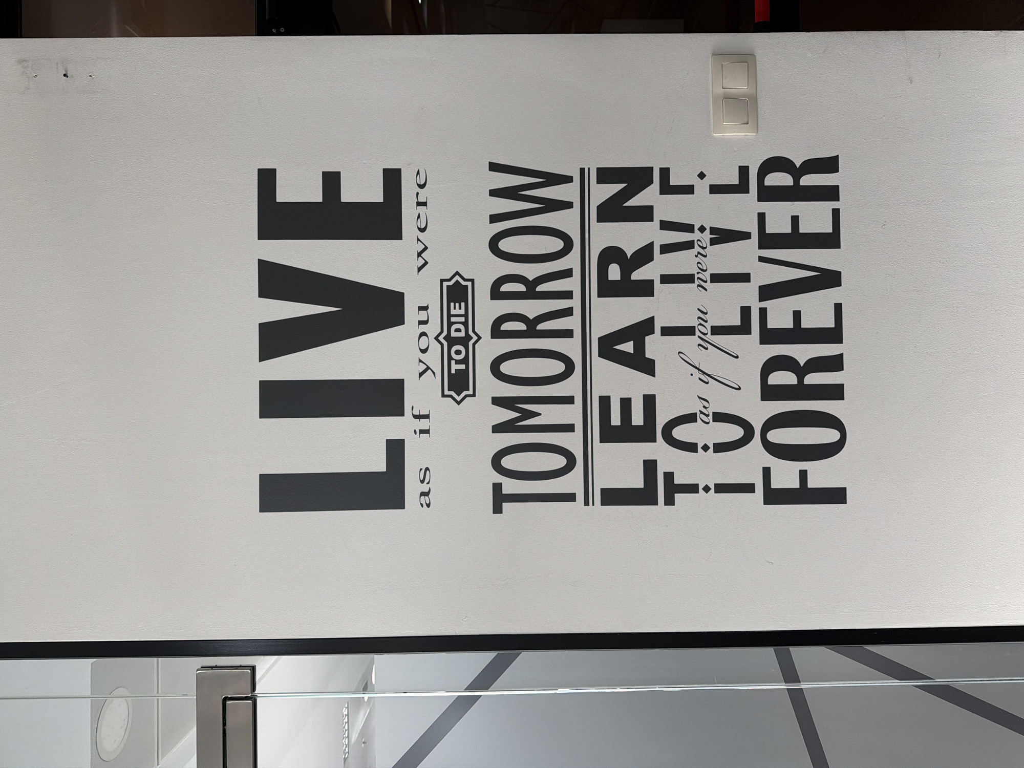 The image shows a close-up of a wall with a motivational quote painted on it in black lettering. The quote reads, "LIVE as if you were TO DIE TOMORROW LEARN as if you were TO LIVE FOREVER." There is a light switch on the upper right part of the wall and a glossy shelf underneath. The location is not clear from this image. I cannot determine the location as Sofia, Bulgaria.
