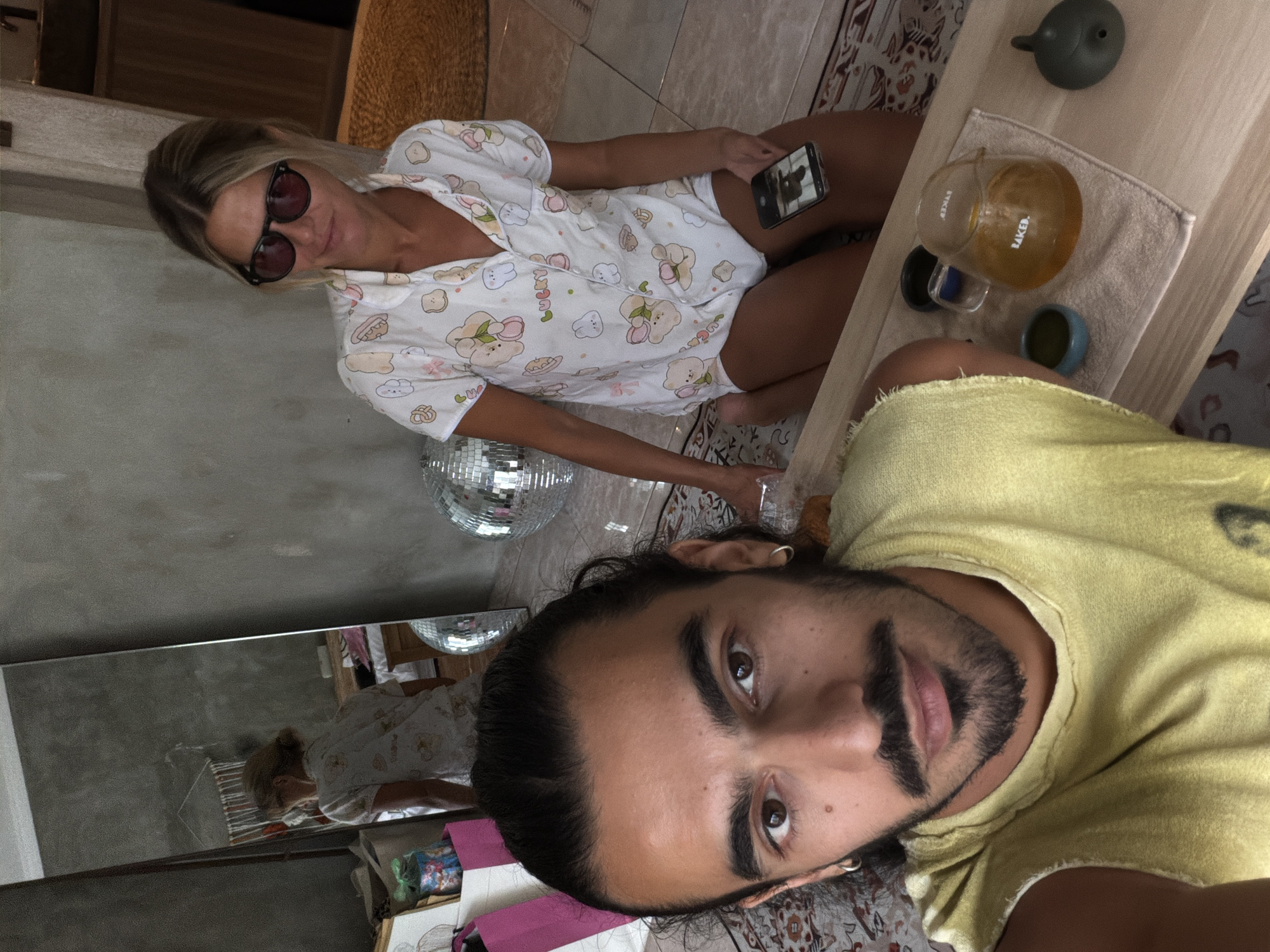 **Description:**
The image shows a man and a woman posing for a selfie indoors, likely in a home or apartment. The man is in the foreground wearing a yellow shirt and has dark hair and a beard. The woman is sitting behind him on the floor. She is wearing a light-colored pajama set with cartoon prints and sunglasses. She is holding a phone in her hand, possibly taking a picture or video.
On a table nearby, there is tea set with a teapot, cup, and small bowls. A disco ball is visible in the background and in the reflection of a mirror. The room is decorated with patterned tiles and rugs.
The location is inferred to be in North Kuta, Indonesia, based on the cultural elements present in the environment and the image metadata.