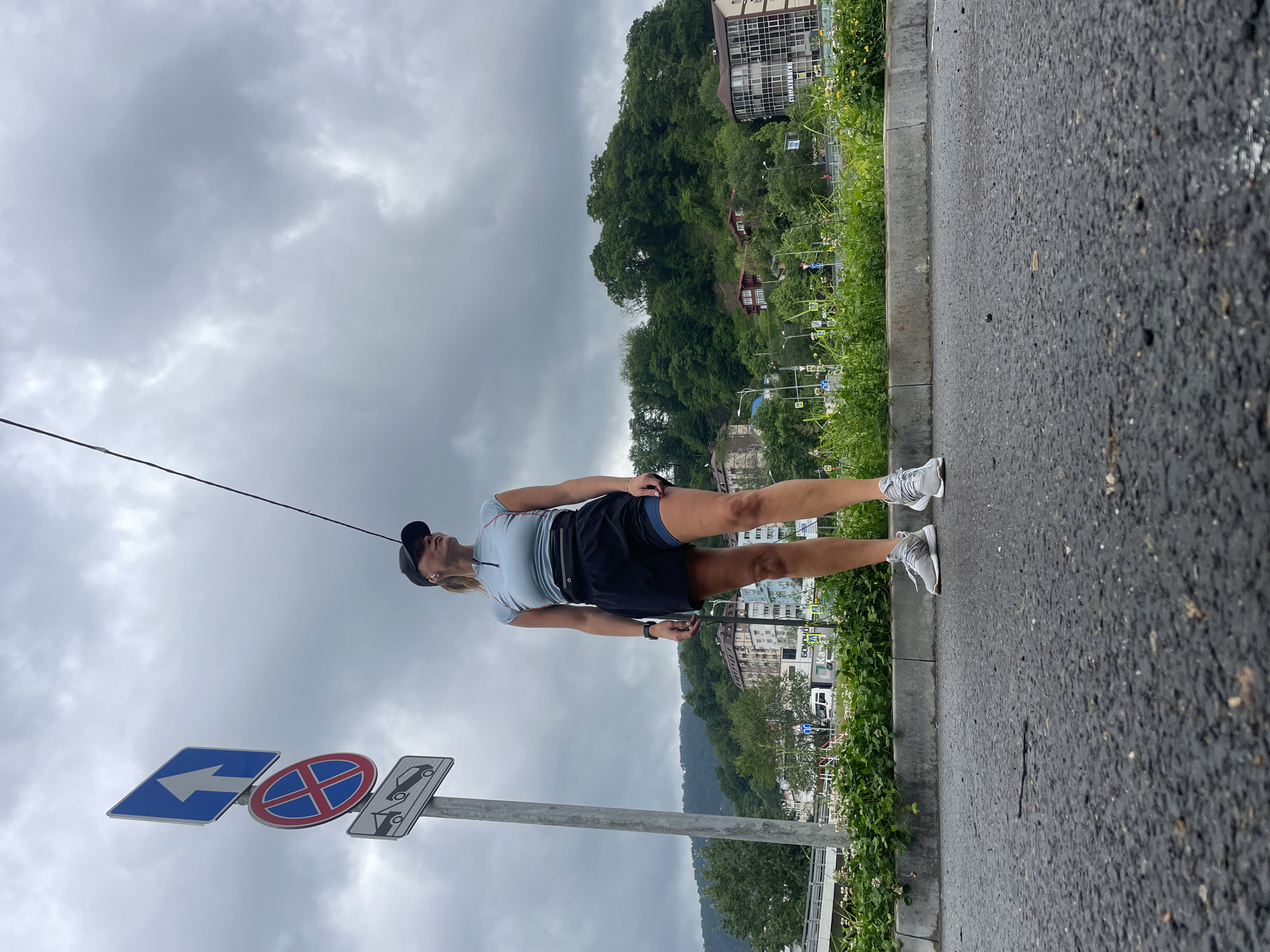 A medium-shot outdoor photograph depicts a fair-skinned woman standing on the sidewalk of a paved road in Sochi, Russia. She wears a baseball cap, a pale-colored short-sleeved shirt, dark shorts, gray sneakers, and a waist bag. The woman stands near a traffic sign composed of three separate signs: one with a blue arrow pointing left, one with a red and blue "no stopping" symbol, and one with a black tow truck symbol. A thin black wire hangs diagonally down from the upper left corner of the frame, passing near the woman's head. The background features dense green foliage and buildings, partly obscured by the trees. Overcast skies fill the upper portion of the frame.
