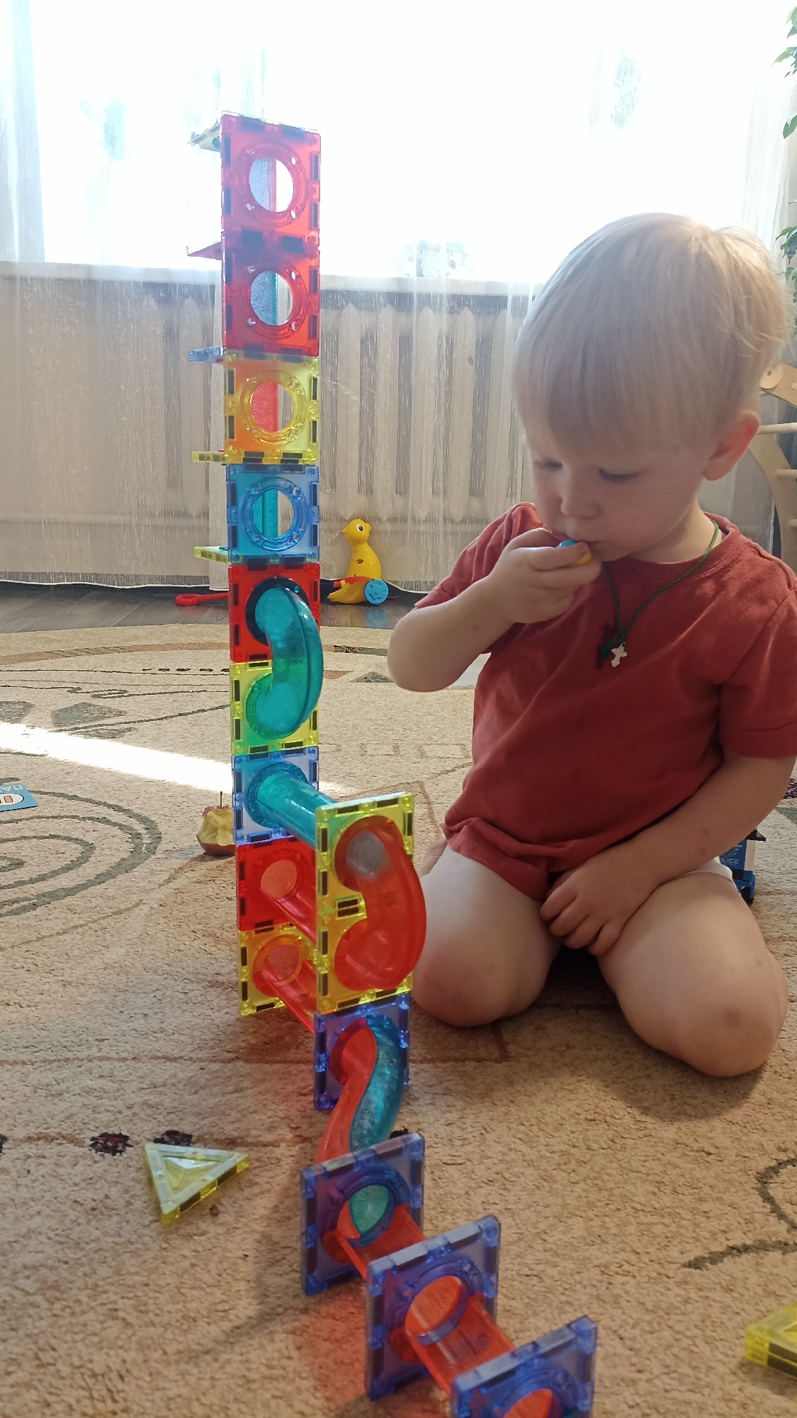 A young blond-haired boy is playing with a marble run on a carpeted floor. He is wearing a red t-shirt and has a green necklace with a cross pendant. The marble run is tall and made of colorful magnetic tiles and translucent tubes. The boy is holding a small, round object, possibly a marble, to his mouth. In the background, there is a window with sheer curtains and a radiator. A yellow toy duck with wheels is visible near the marble run. The lighting suggests it is daytime. An apple core is on the floor near the marble run, and a yellow triangular tile is also visible on the carpet. The scene appears to be in a home.