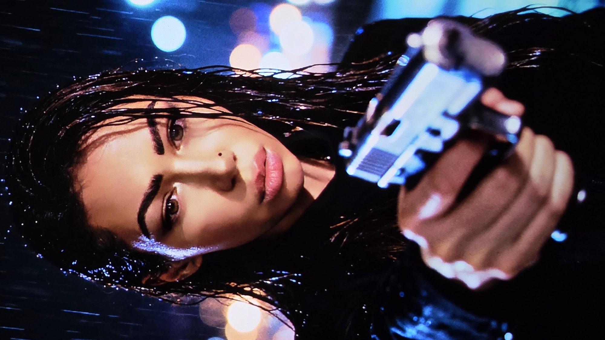 A woman lies on her side in the rain, holding a silver pistol. Her hair is dark and wet, slicked back from her face. She wears a black top. The background features blurry lights, suggesting a city setting. The scene does not provide enough data to conclude the location is London.